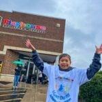 A child in an event T-shirt raises both arms in celebration outside the BDI Playhouse building