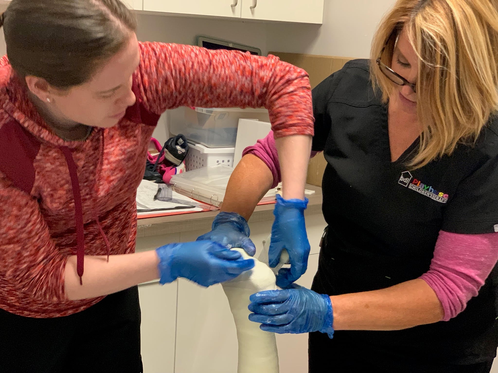 Two therapists wearing blue gloves apply a cast to a child's lower leg