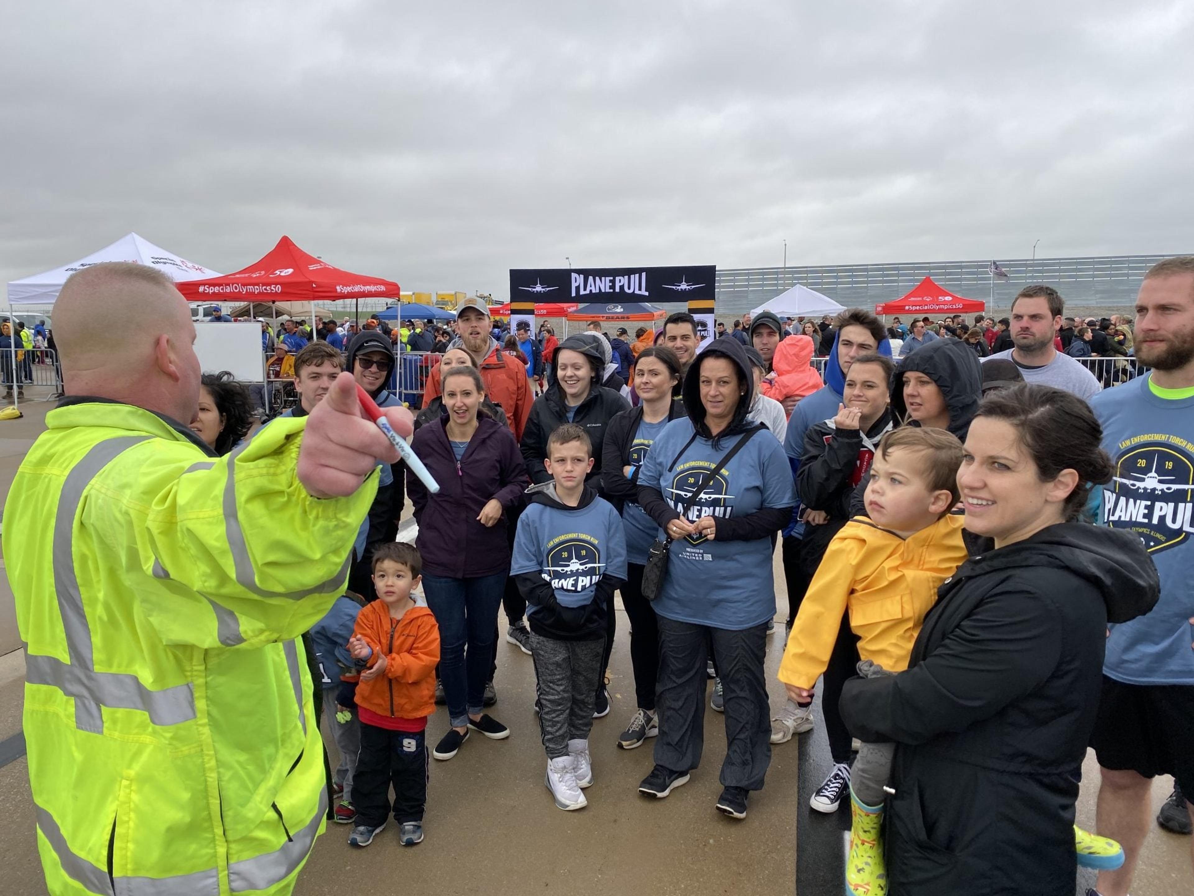 Staff and families gather on the tarmac at the Plane Pull event as a worker in a safety jacket gives directions