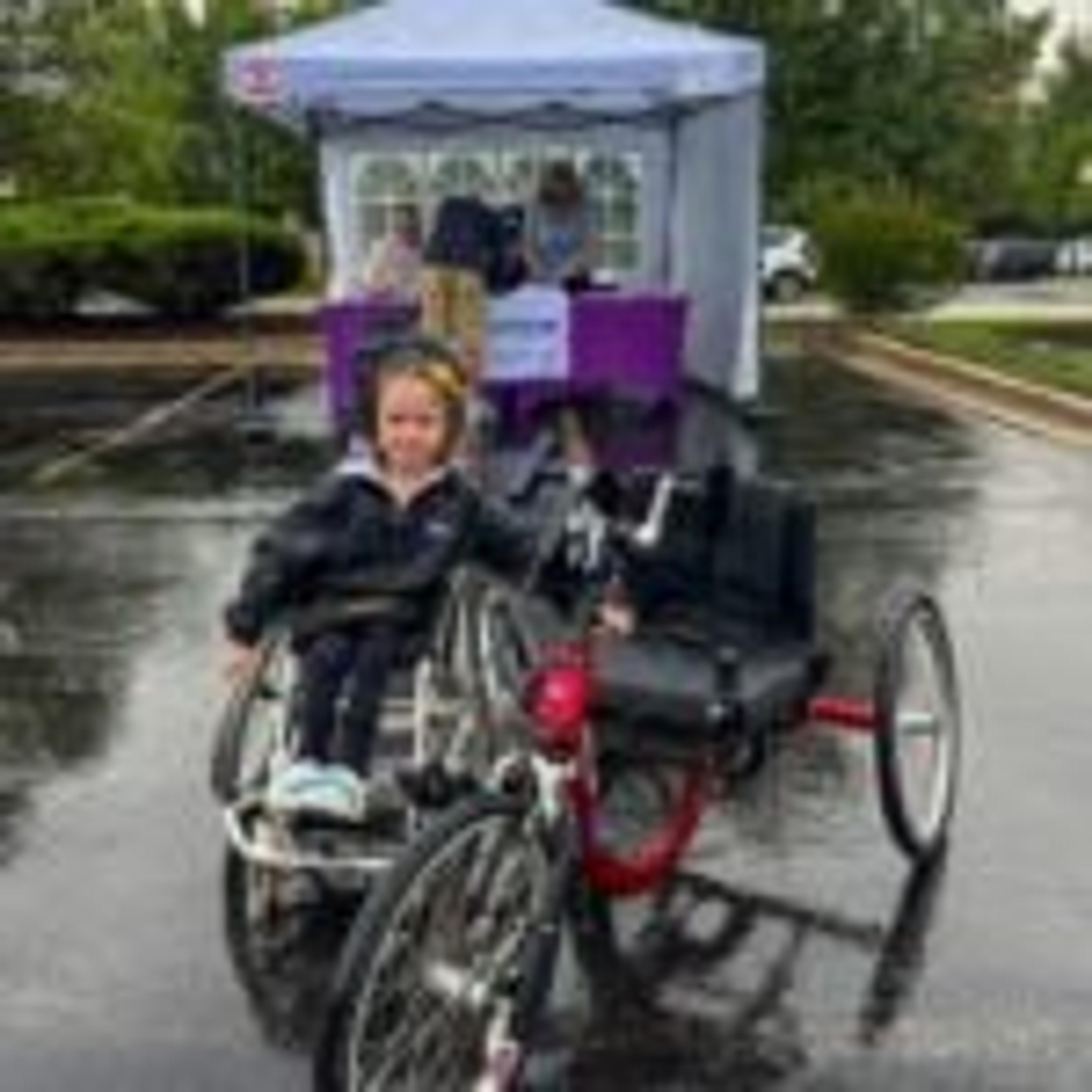 A smiling child with an adaptive tricycle on a rain-soaked parking lot, an event tent in the background