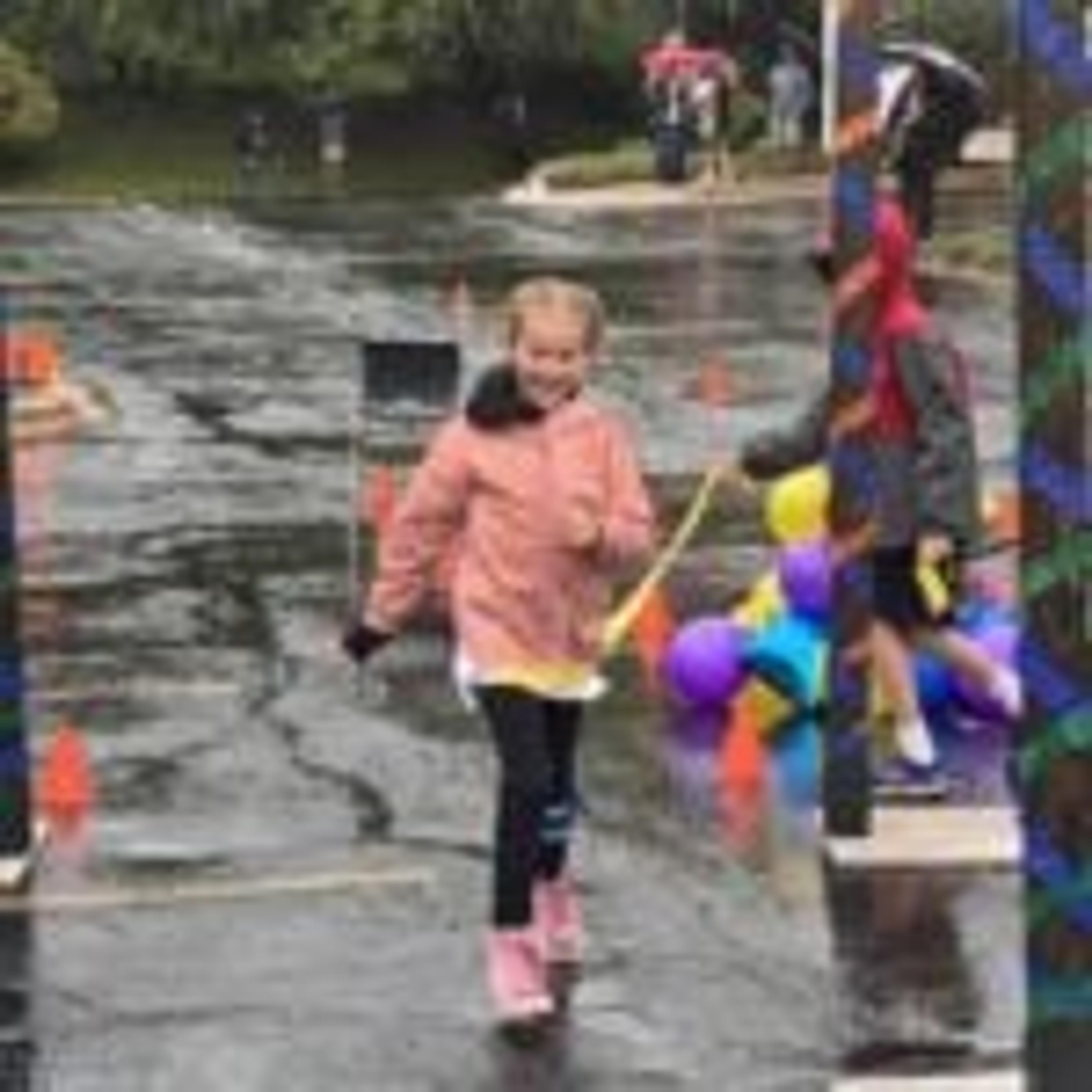 A smiling girl in a pink jacket runs along the wet course past balloons and decorated poles