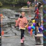 A smiling girl in a pink jacket runs along the wet course past balloons and decorated poles