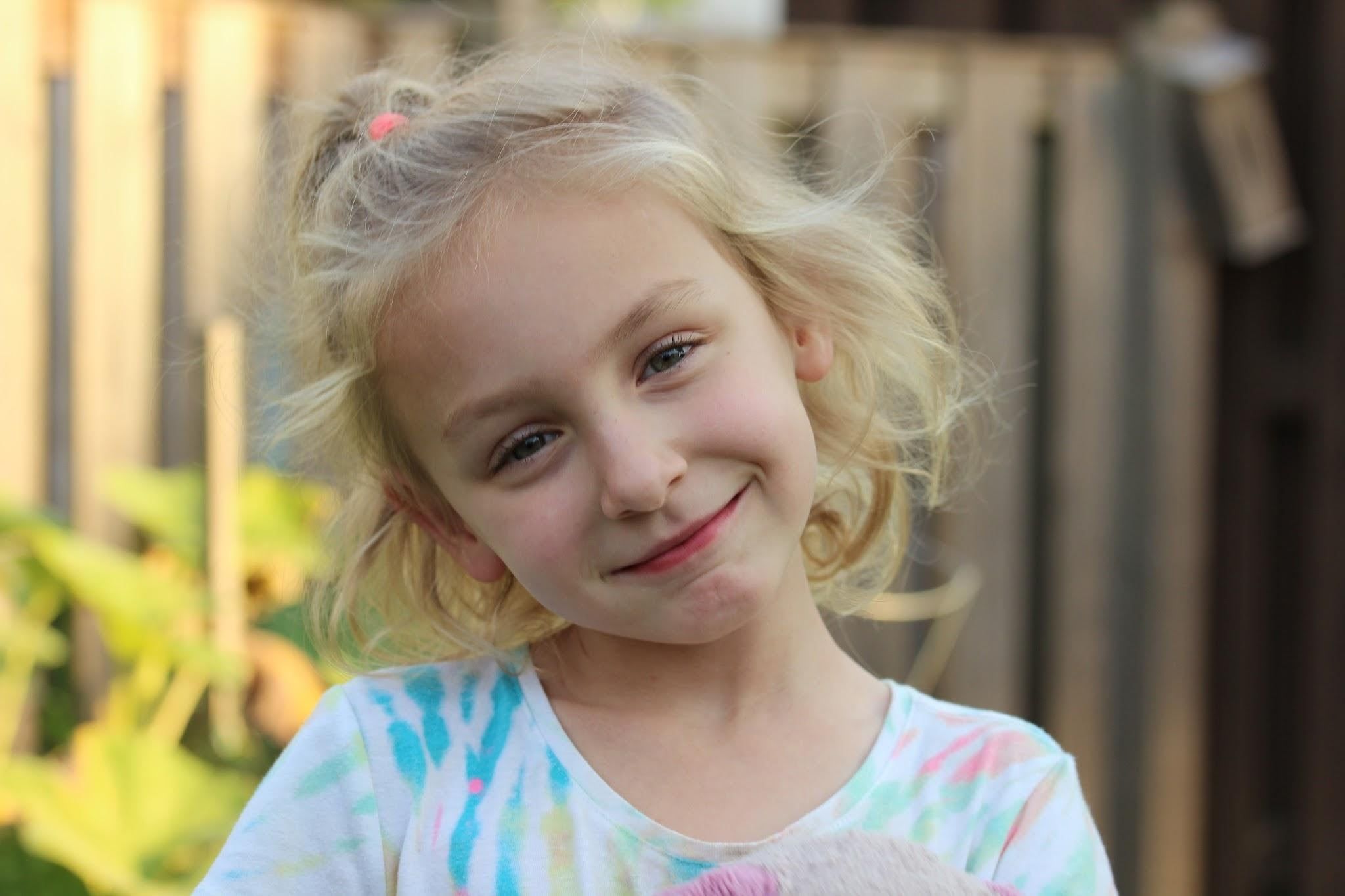 A smiling young girl with curly blonde hair tilts her head, standing outdoors by a wooden fence