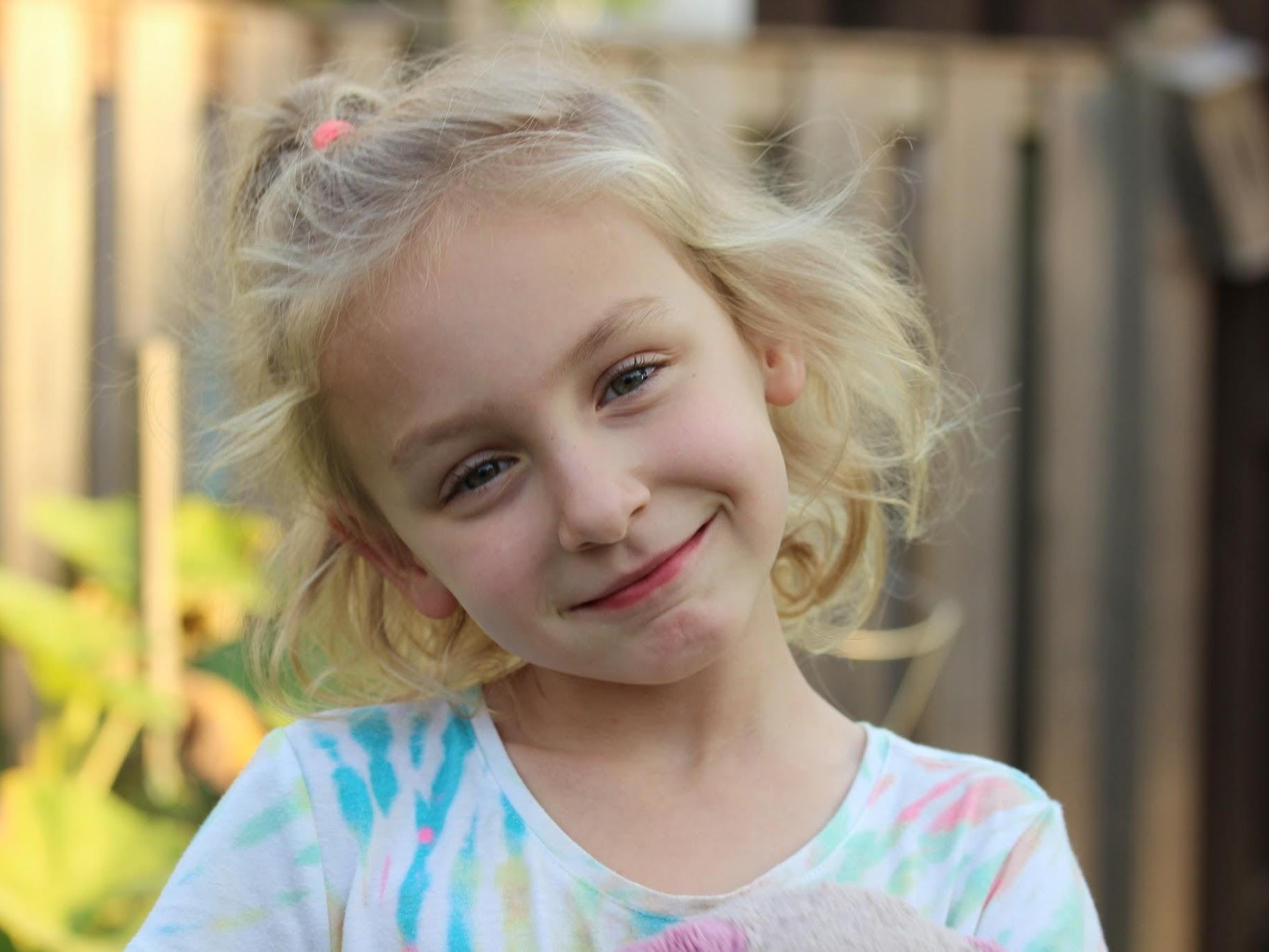 A smiling young girl with curly blonde hair tilts her head, standing outdoors by a wooden fence