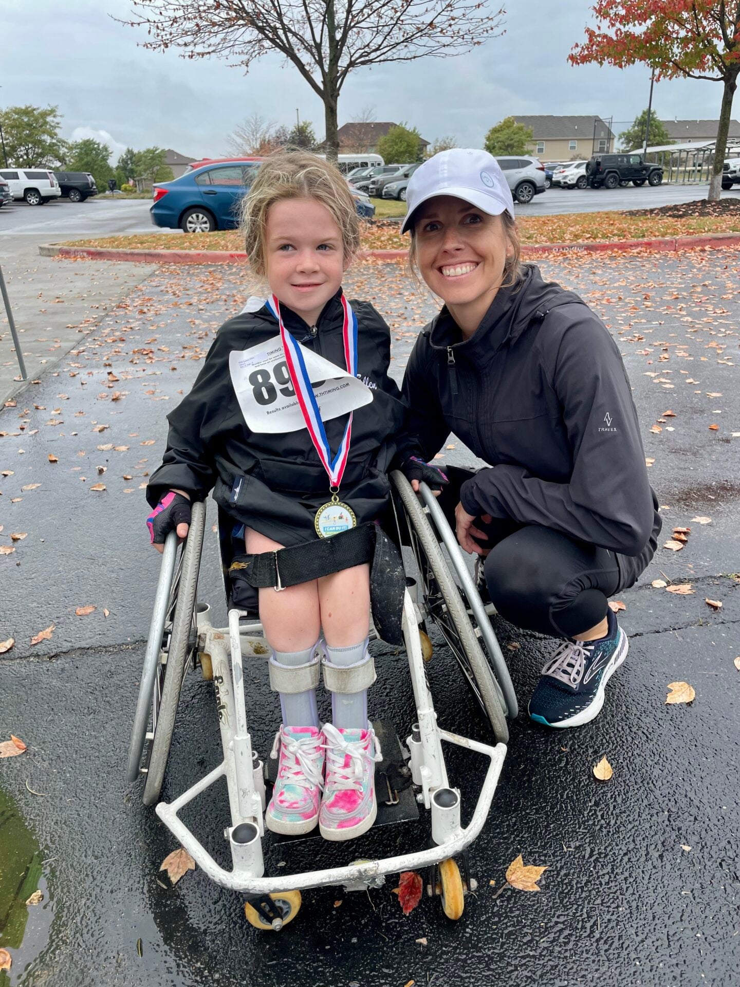 A girl wearing a race bib and medal stands in a walker beside a smiling woman crouching next to her in a parking lot