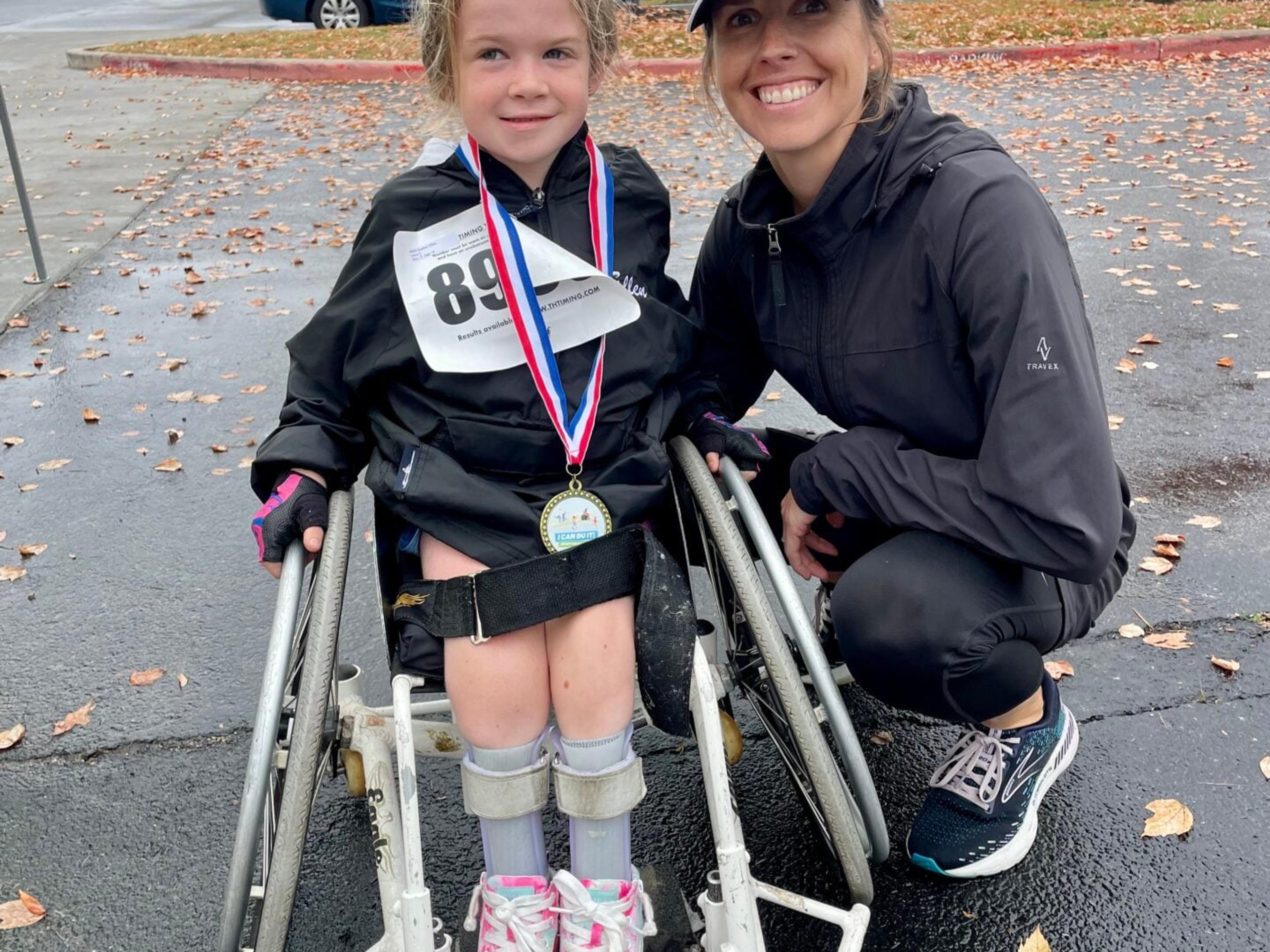A girl wearing a race bib and medal stands in a walker beside a smiling woman crouching next to her in a parking lot