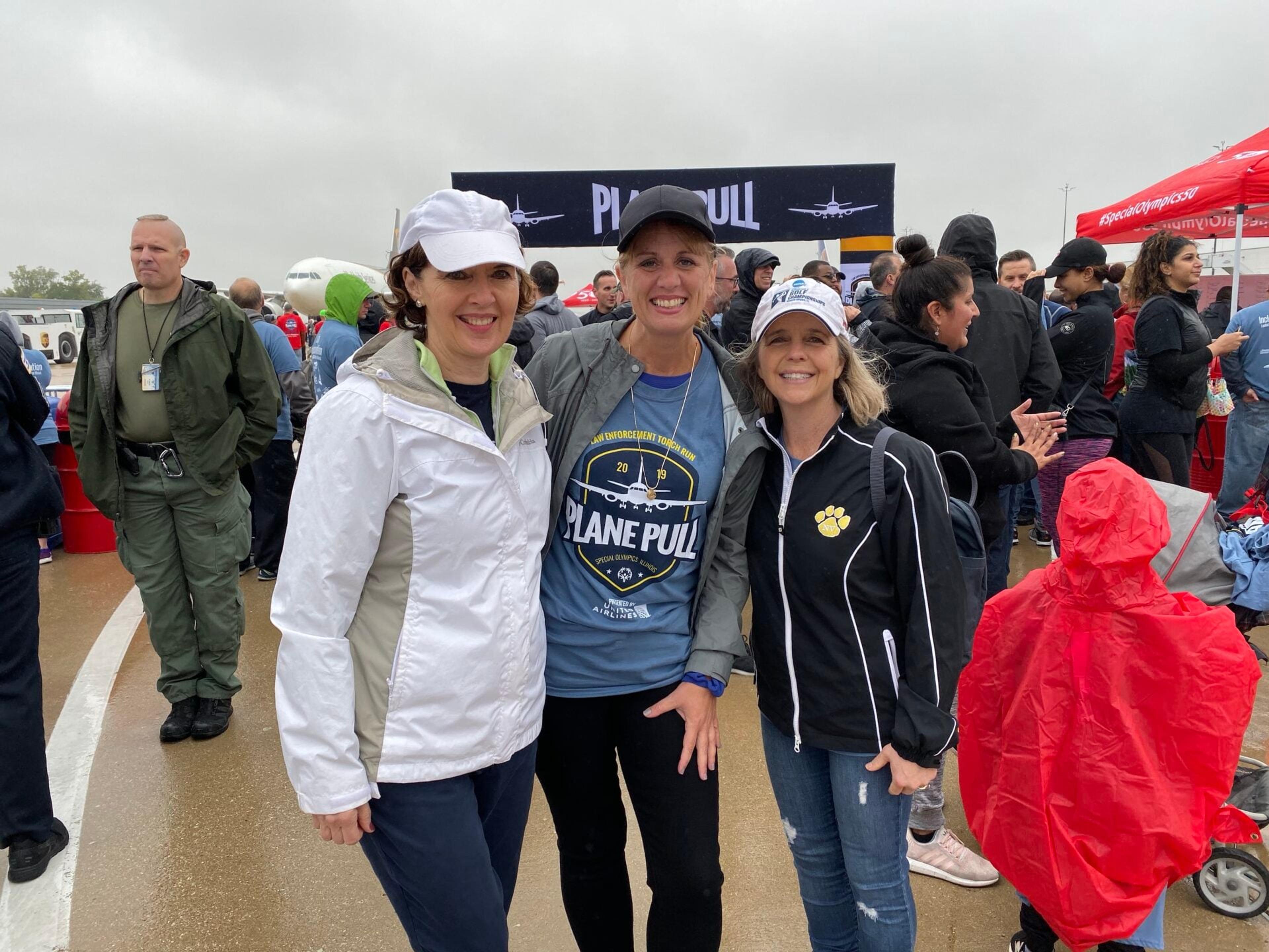 Three staff members in caps and jackets pose together at the Plane Pull event on a rainy day