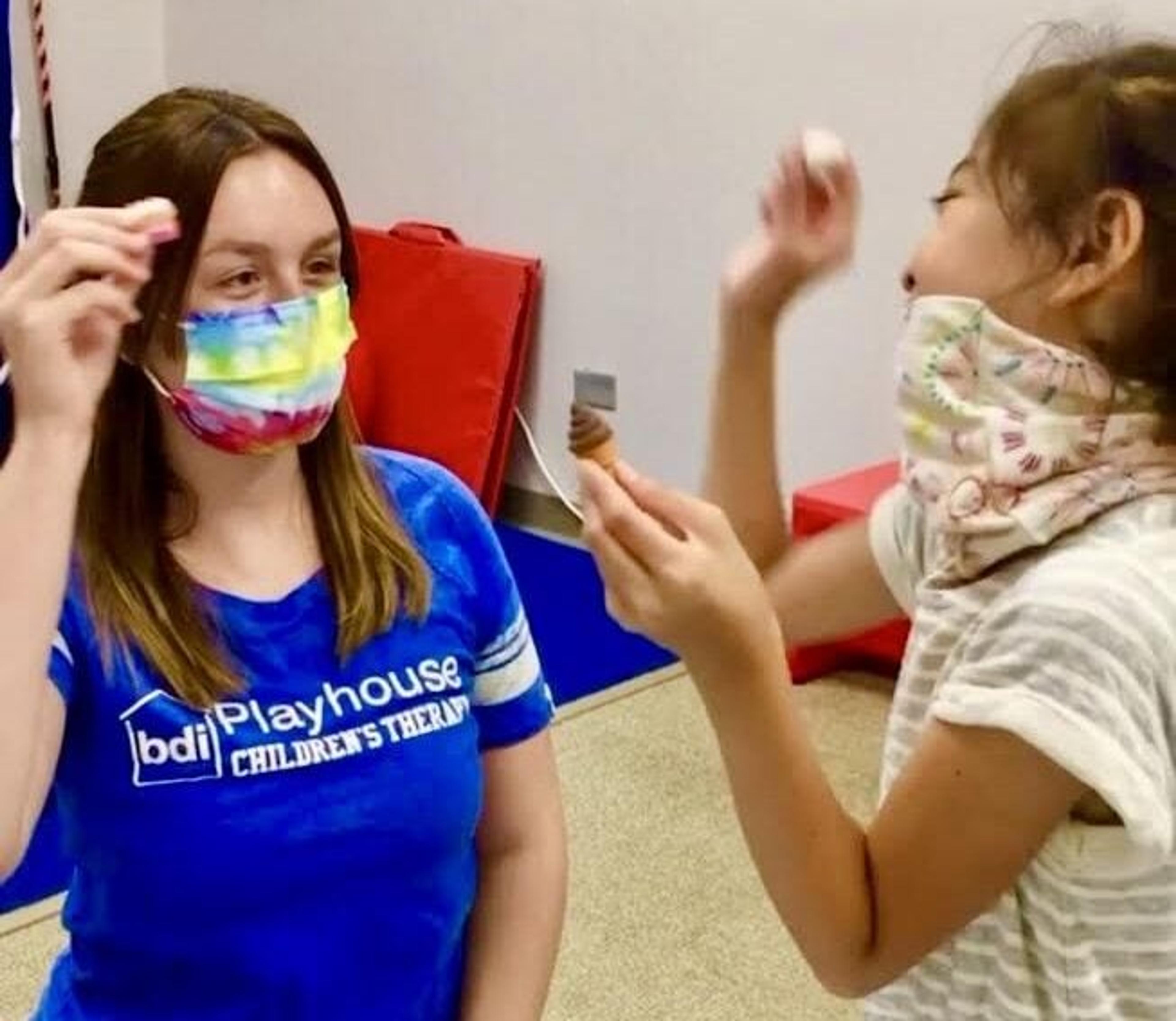 A team member in a BDI Playhouse shirt and a girl, both wearing face masks, play with toy ice cream cones in the gym