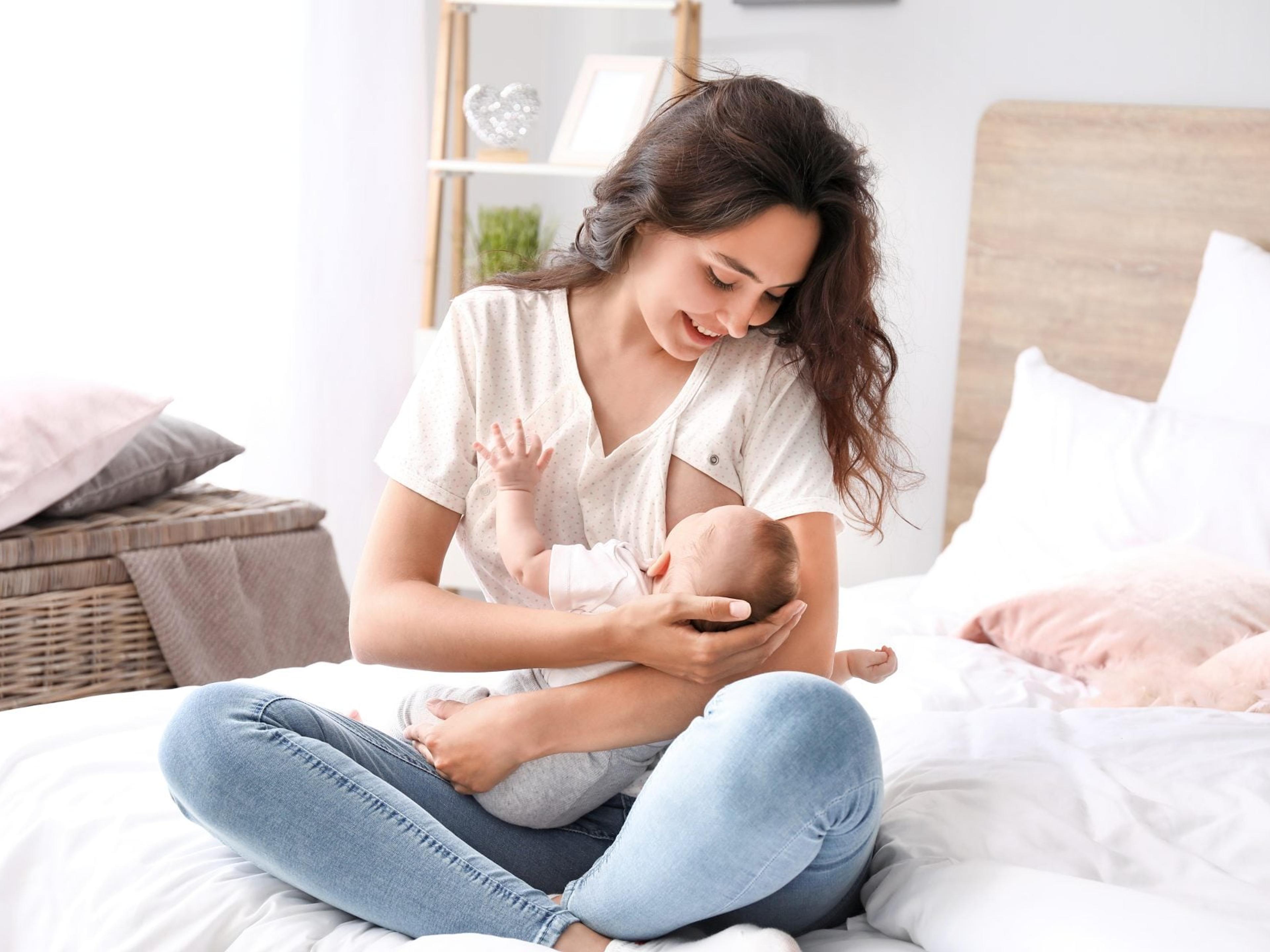 A smiling mother sits cross-legged on a bed breastfeeding her baby