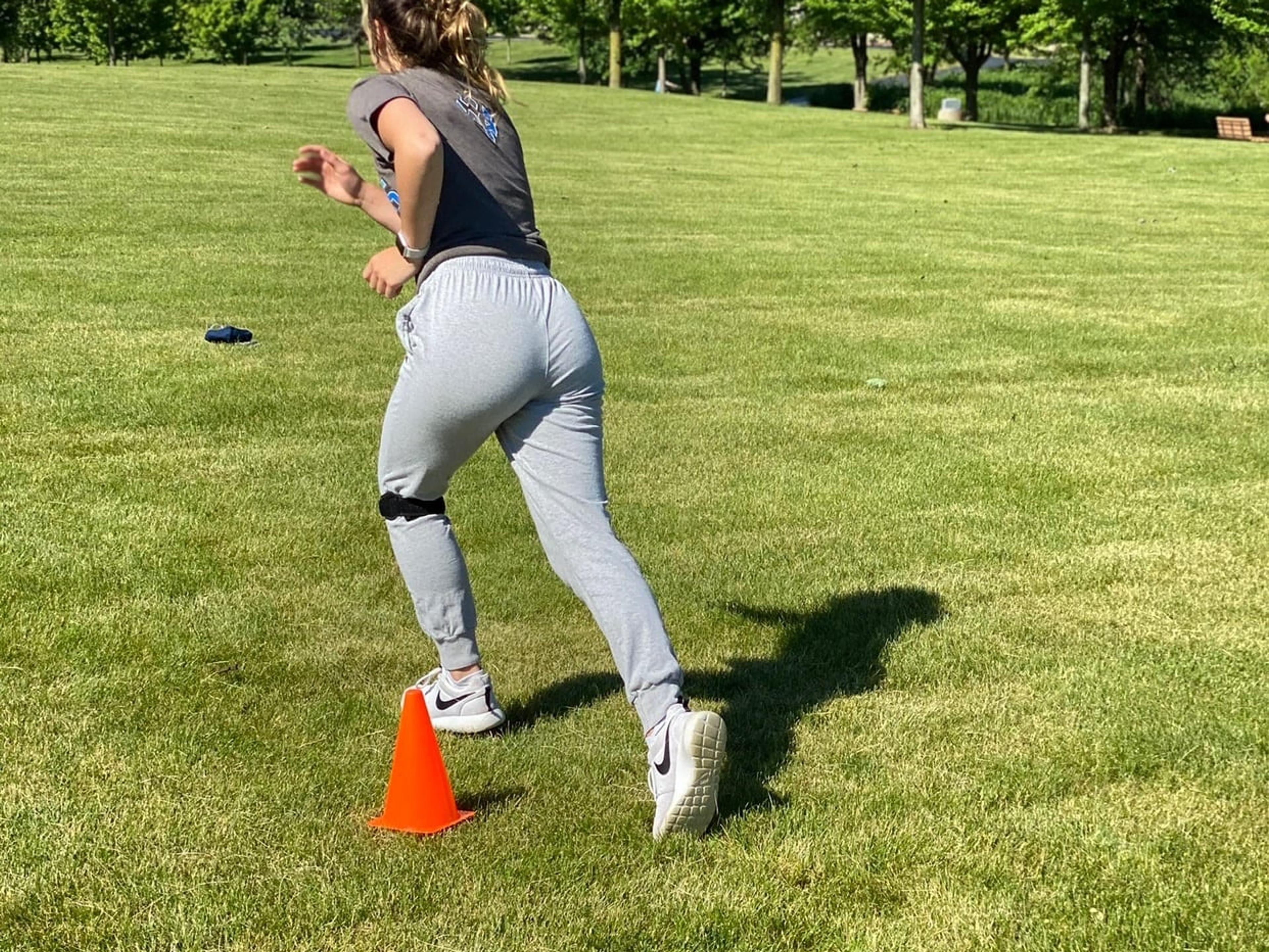 A teen athlete wearing a knee strap pushes off to sprint past an orange cone on a grass field