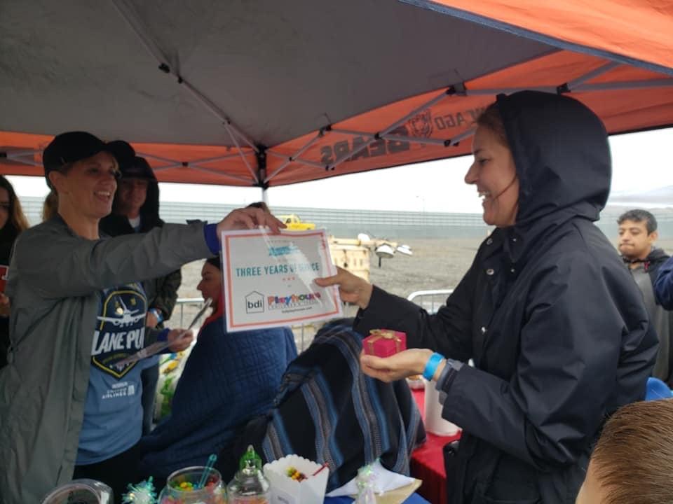 A staff member presents a BDI Playhouse three-year service certificate to a smiling colleague under an event tent