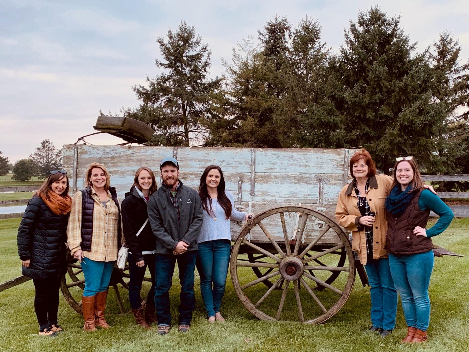 Seven staff members pose beside an old wooden wagon on a staff barn day outing