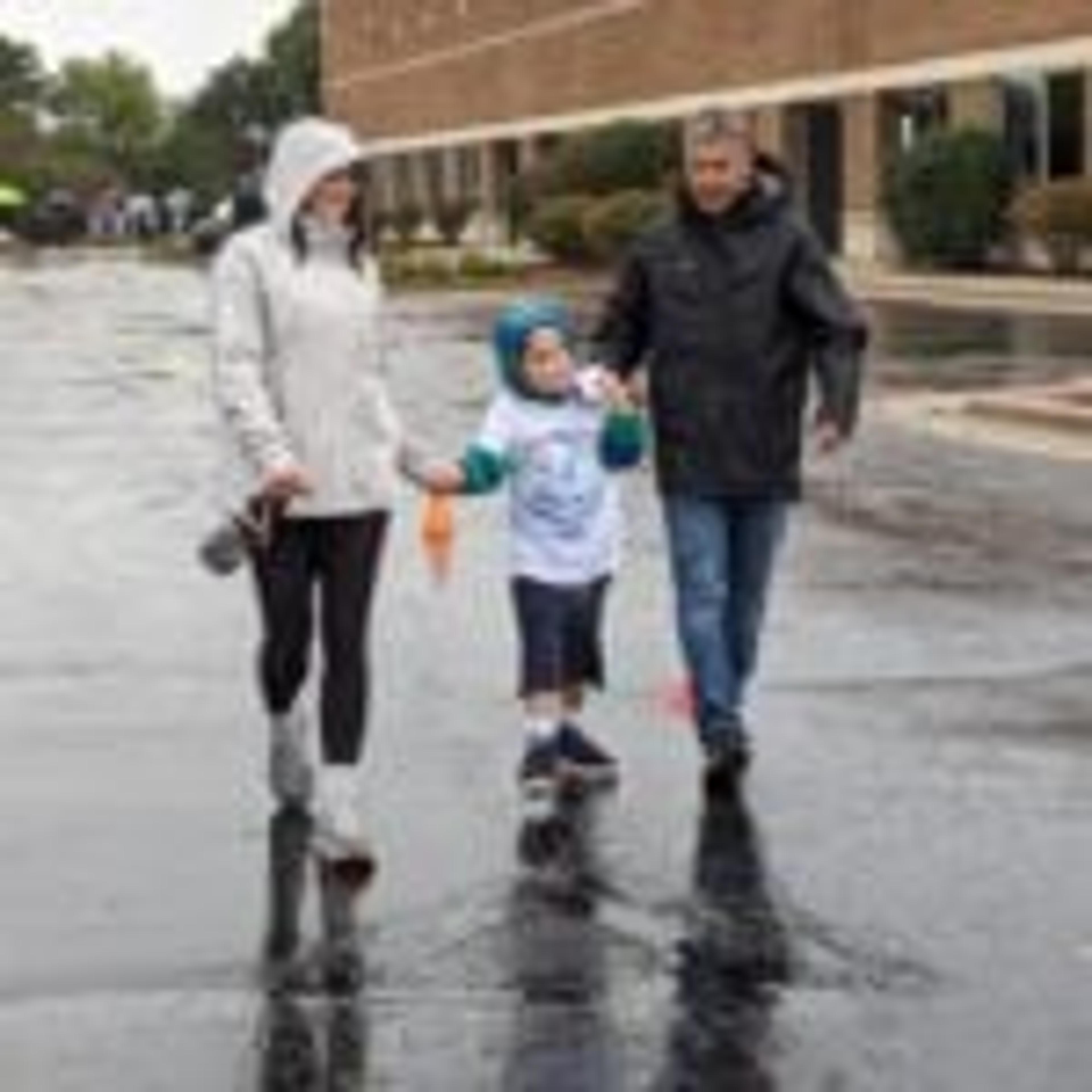 Two adults in rain jackets hold a young child's hands as they walk the wet course together