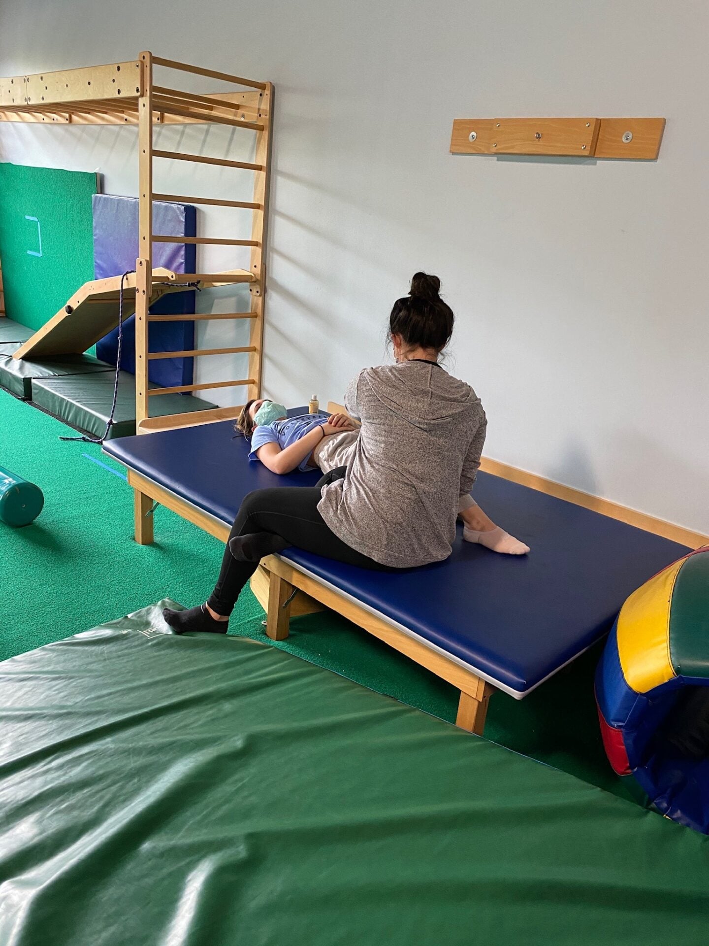 A therapist sits on a padded treatment table working with a patient who lies on their back in the therapy gym