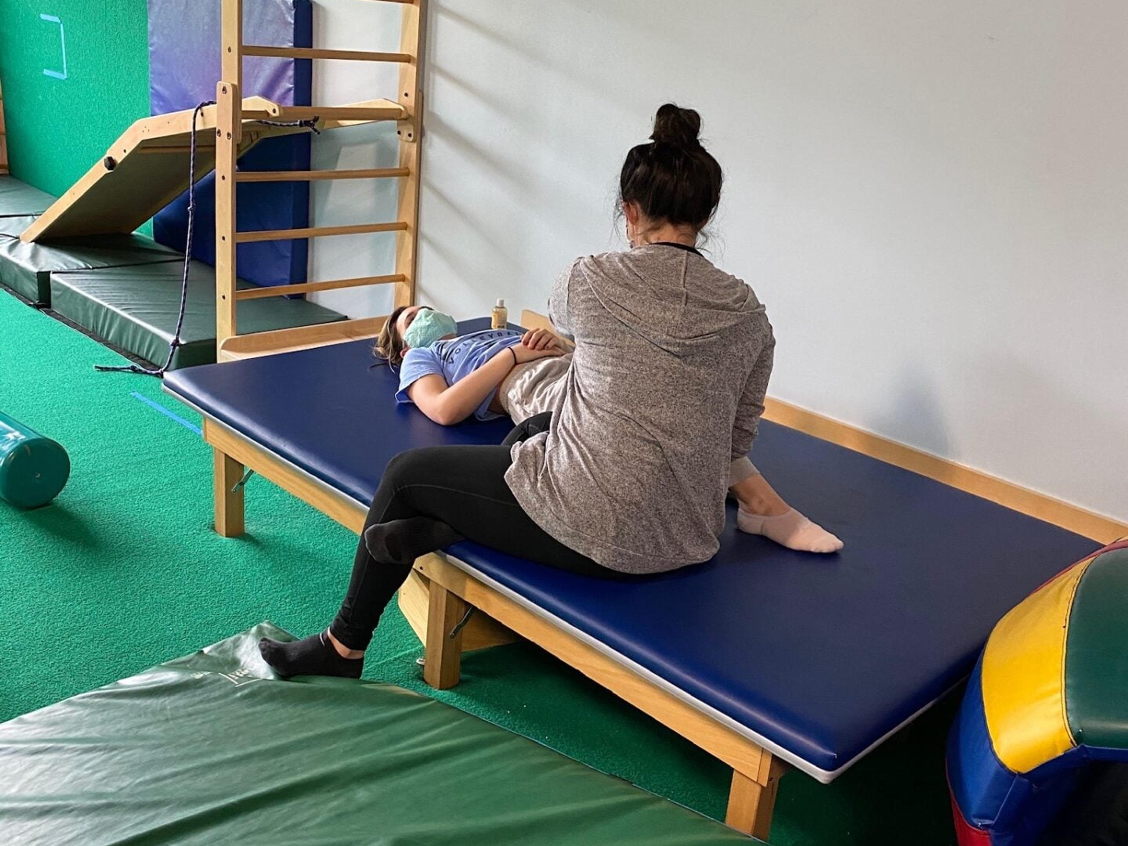 A therapist sits on a padded treatment table working with a patient who lies on their back in the therapy gym