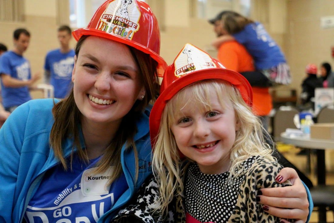 A smiling team member and a young girl, both wearing red toy firefighter hats, pose together at a community event