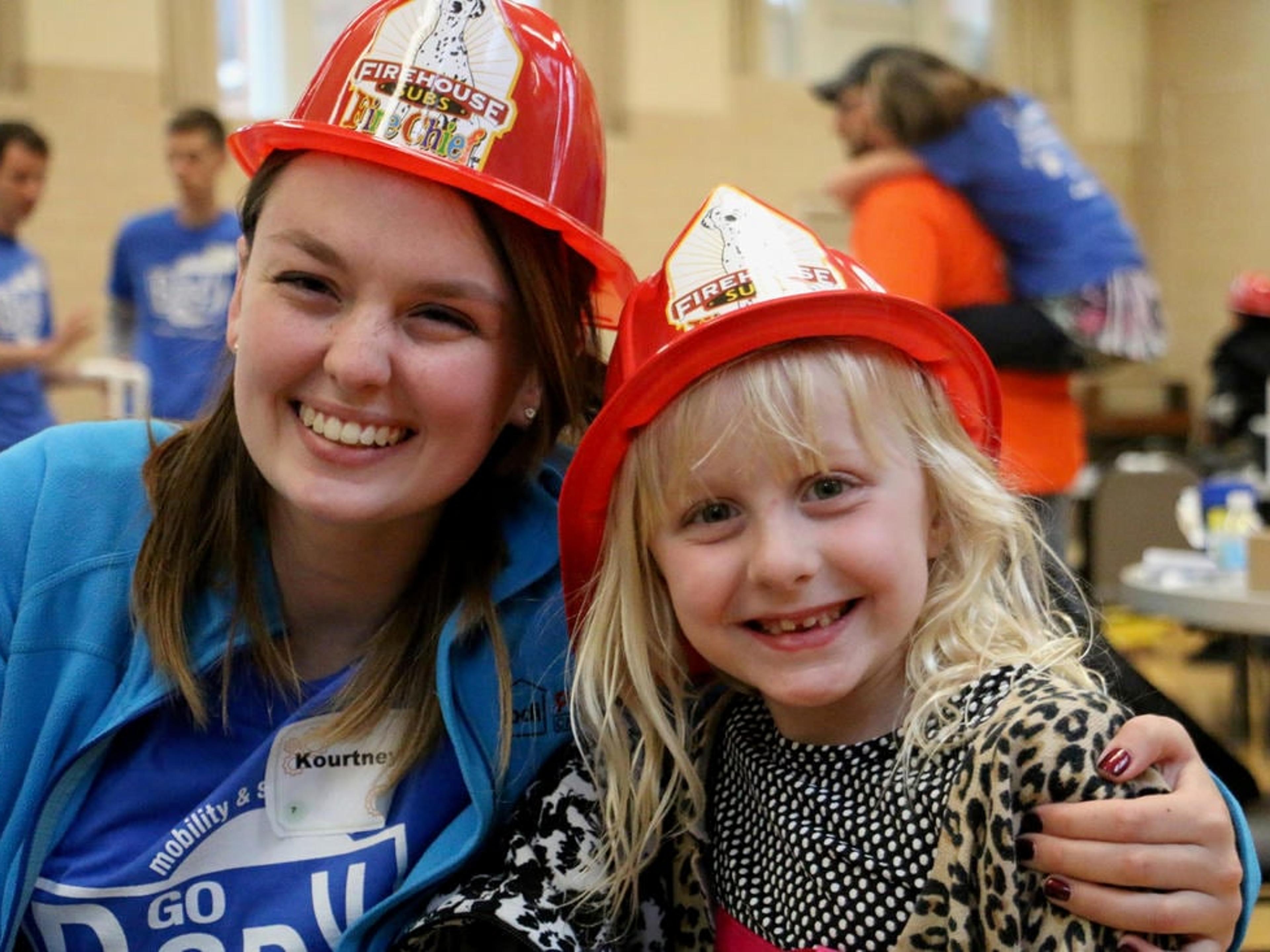 A smiling team member and a young girl, both wearing red toy firefighter hats, pose together at a community event