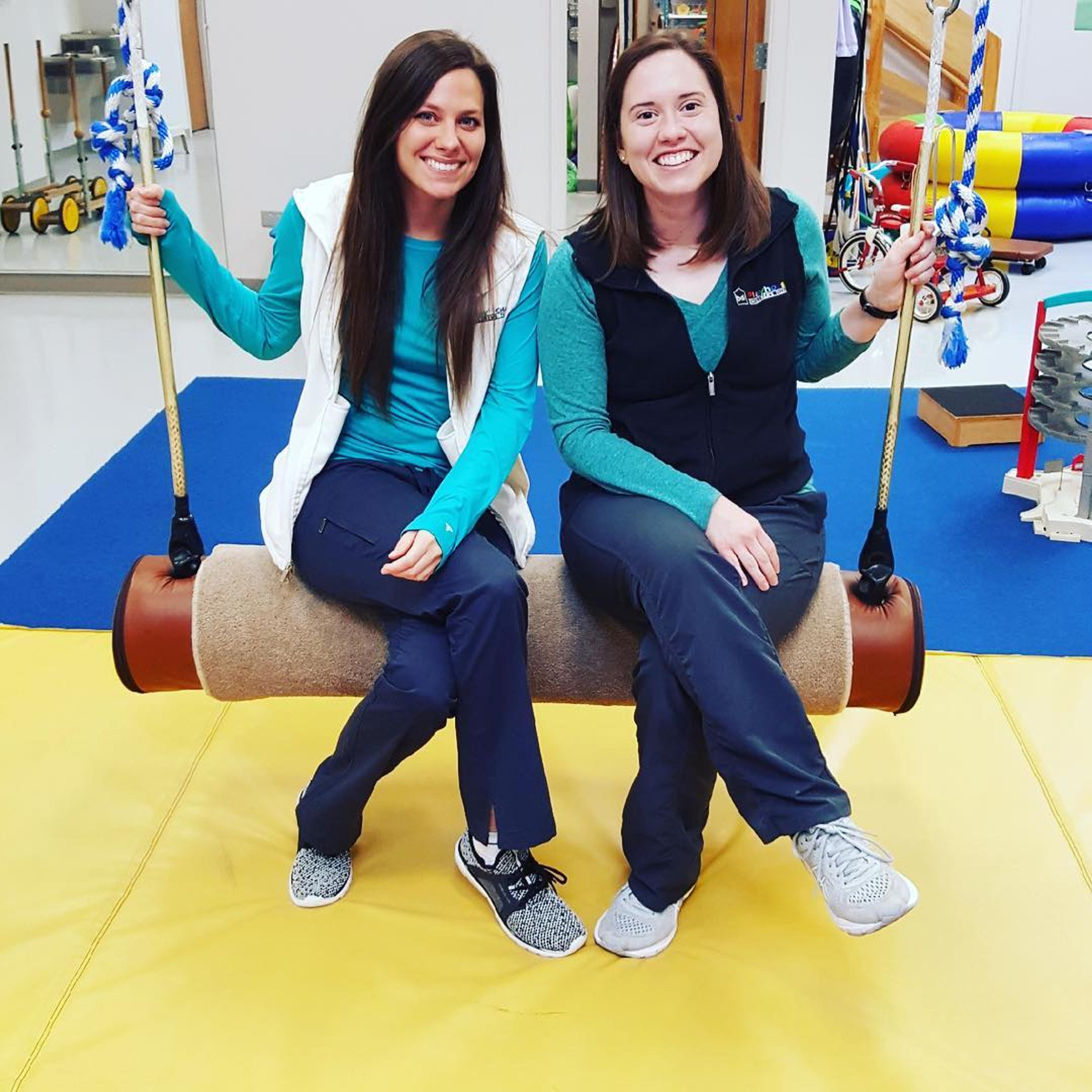 Two smiling therapists sit side by side on a bolster swing in the therapy gym