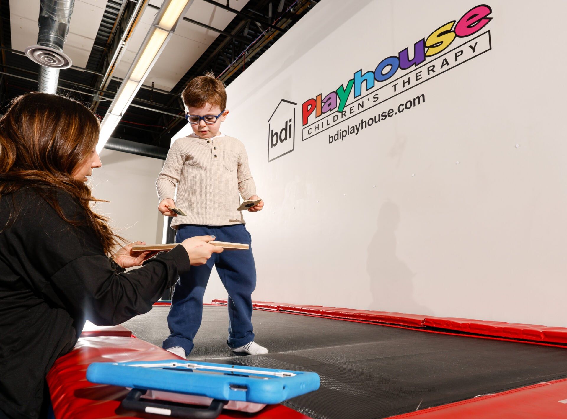 A therapist hands cards to a boy in glasses standing on a trampoline beneath the BDI Playhouse wall sign