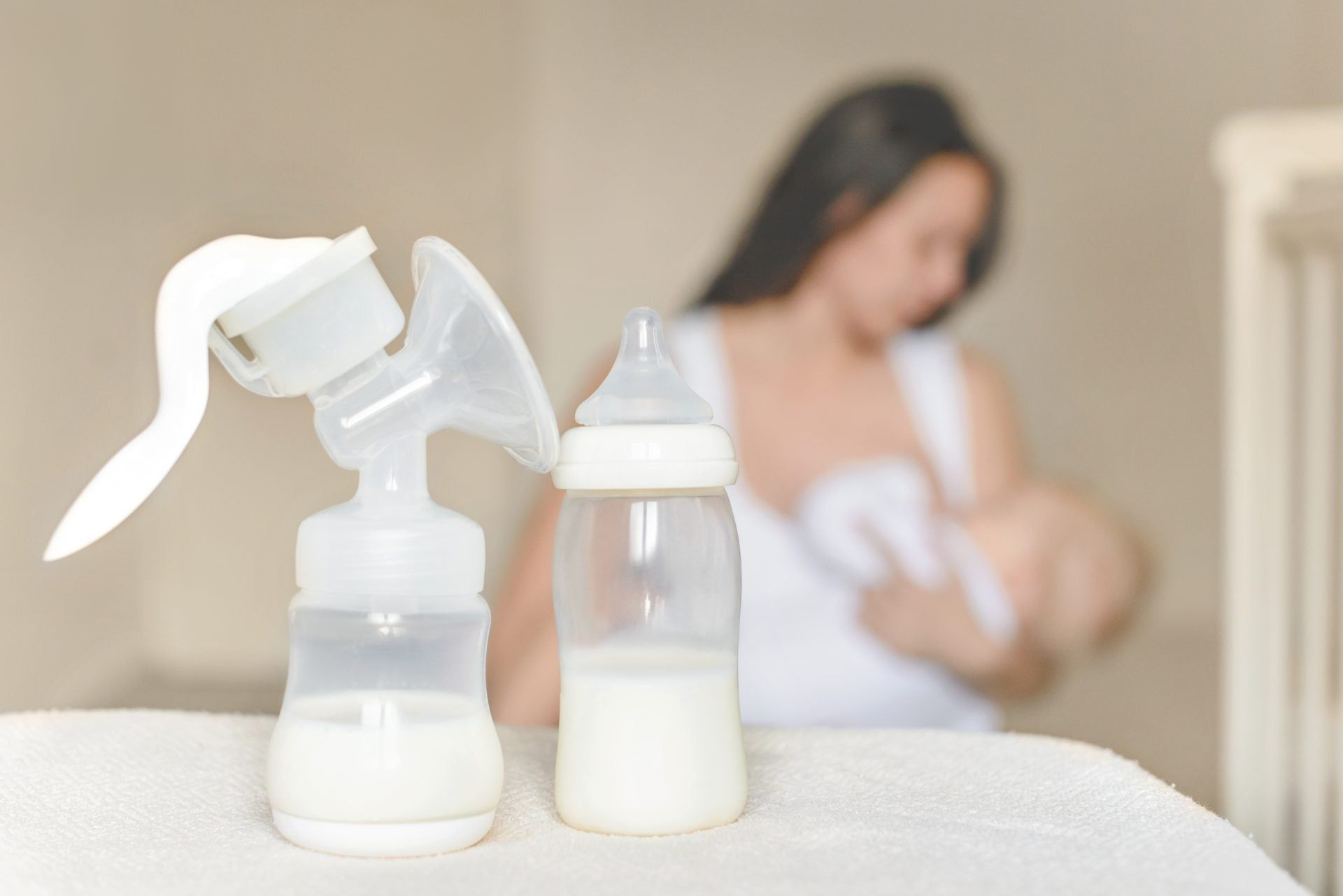 A breast pump and a bottle of milk on a table, with a mother nursing her baby softly blurred in the background