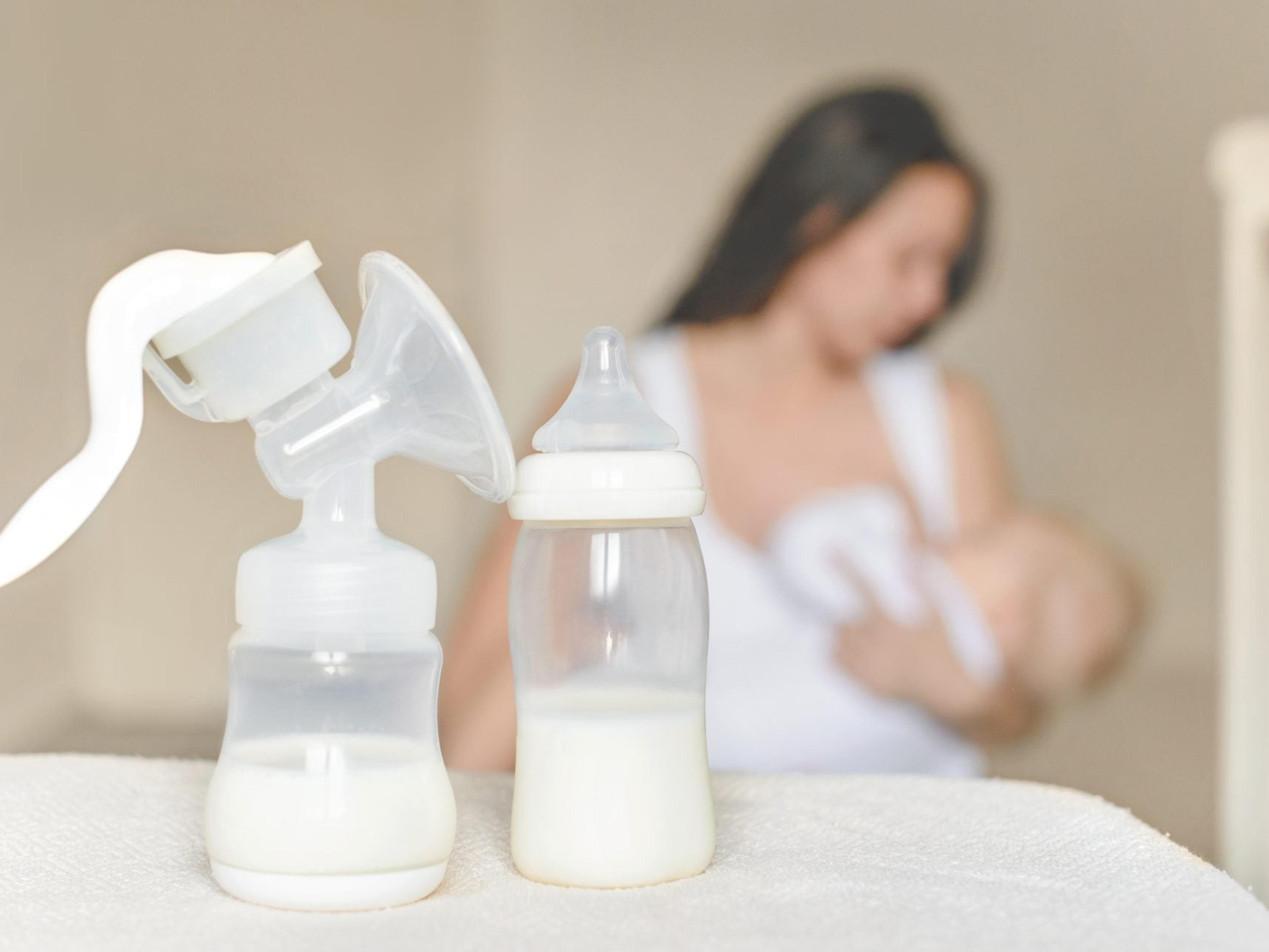 A breast pump and a bottle of milk on a table, with a mother nursing her baby softly blurred in the background
