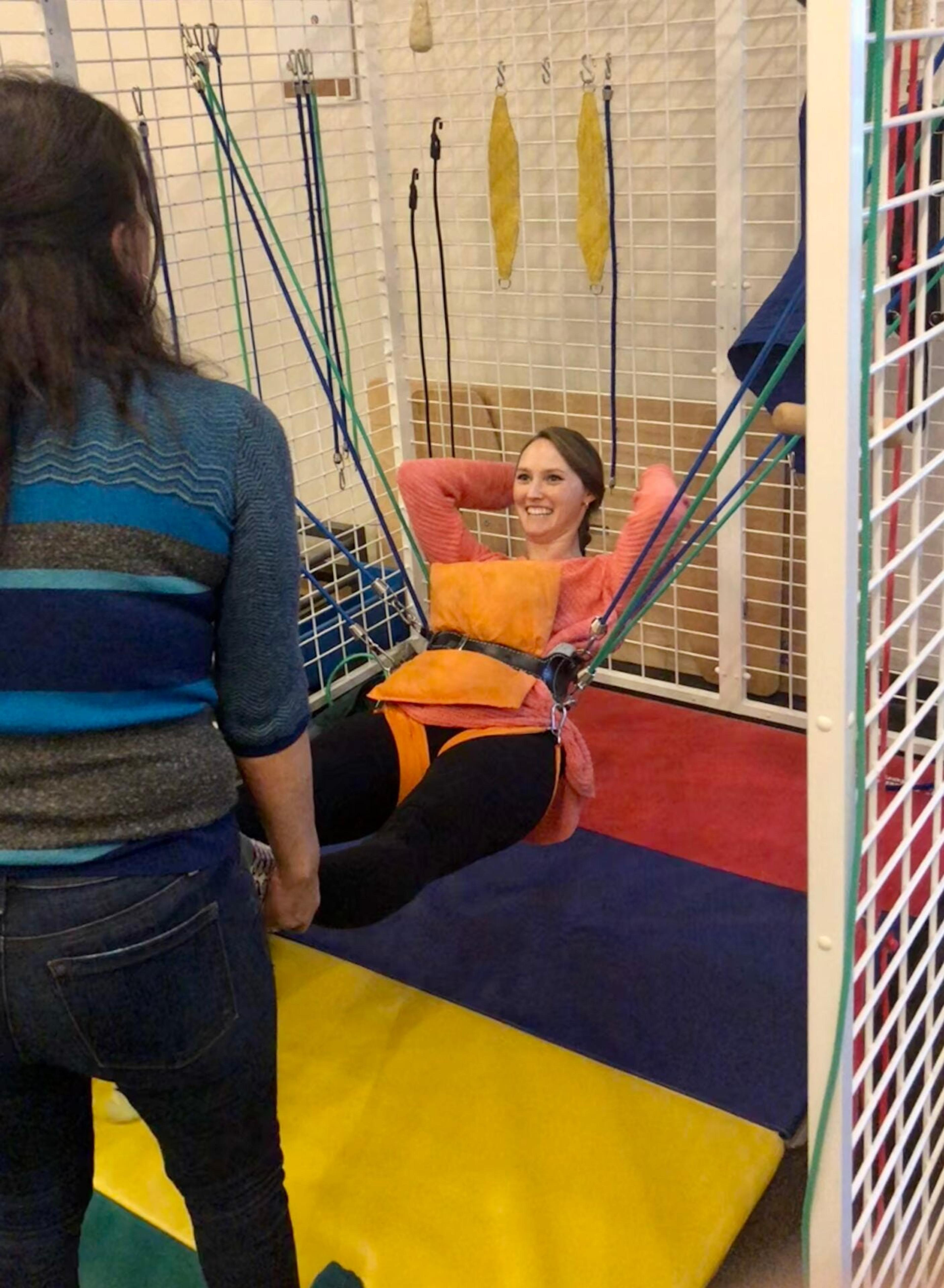 A smiling therapist reclines in a harness suspended by bungee cords inside a therapy cage during staff training
