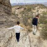 A child and an adult with a backpack hike along a rocky desert trail