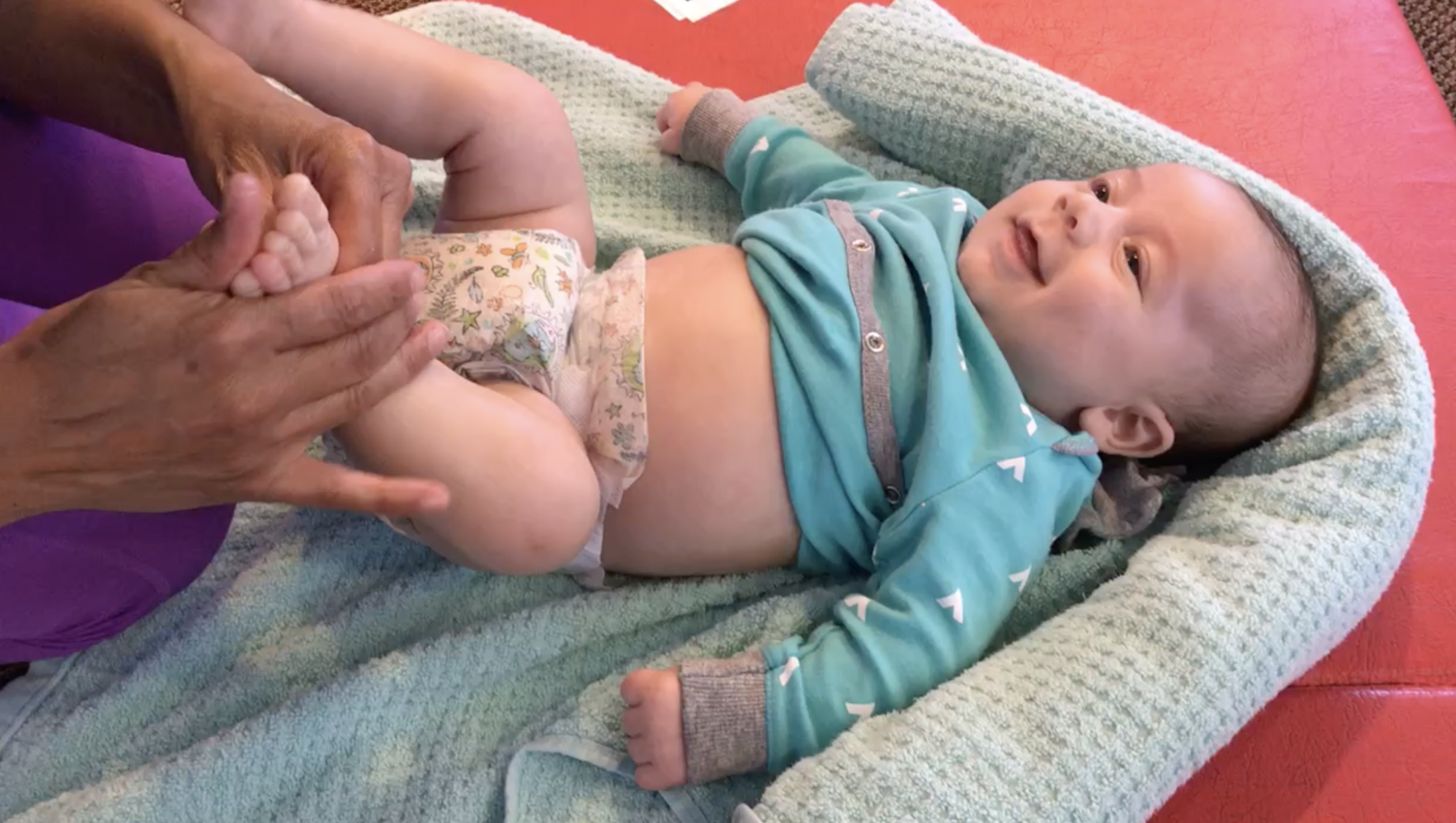 An adult gently holds a smiling baby's feet as the baby lies in a padded lounger on a mat