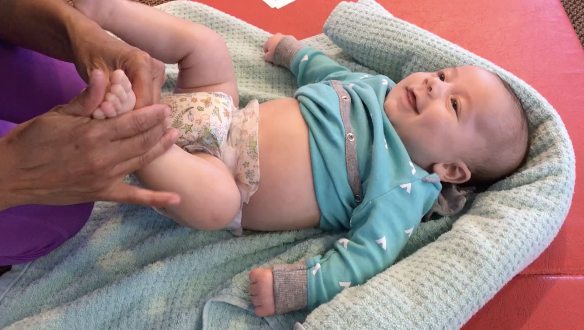 An adult gently holds a smiling baby's feet as the baby lies in a padded lounger on a mat