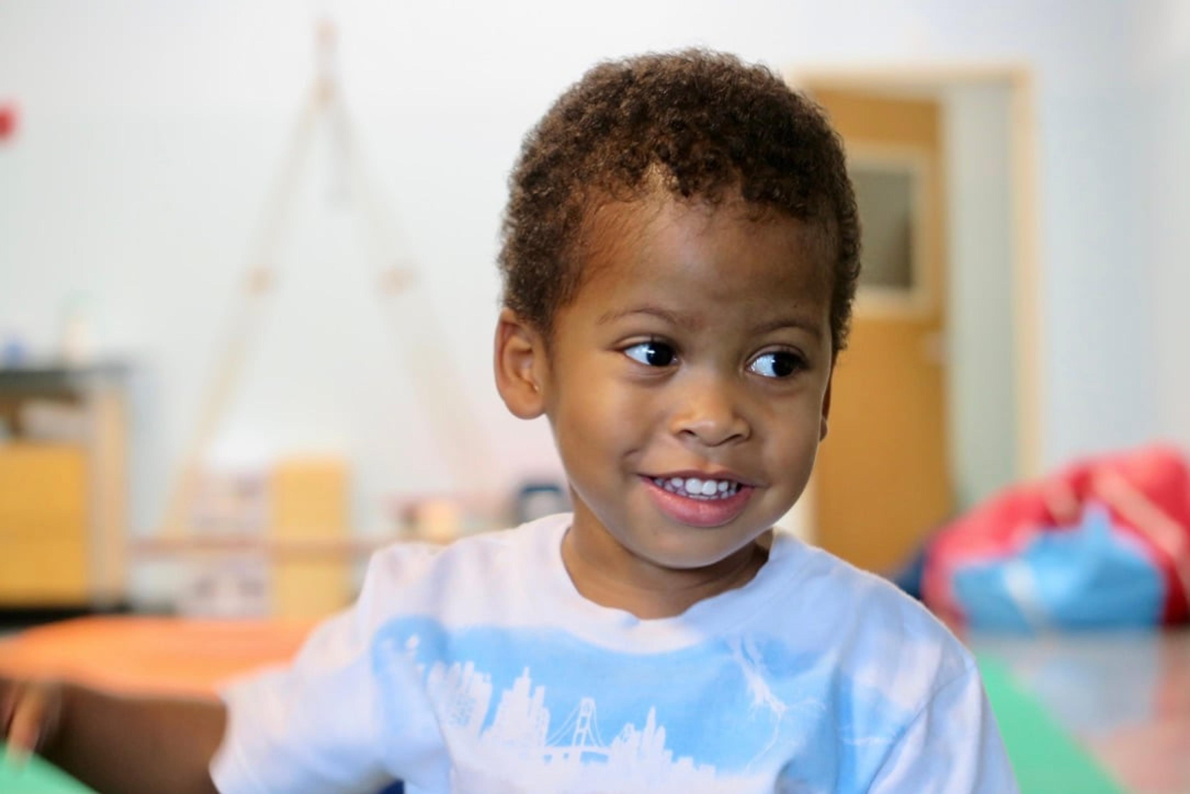 A smiling young boy in a tie-dye shirt looks off to the side in a playroom