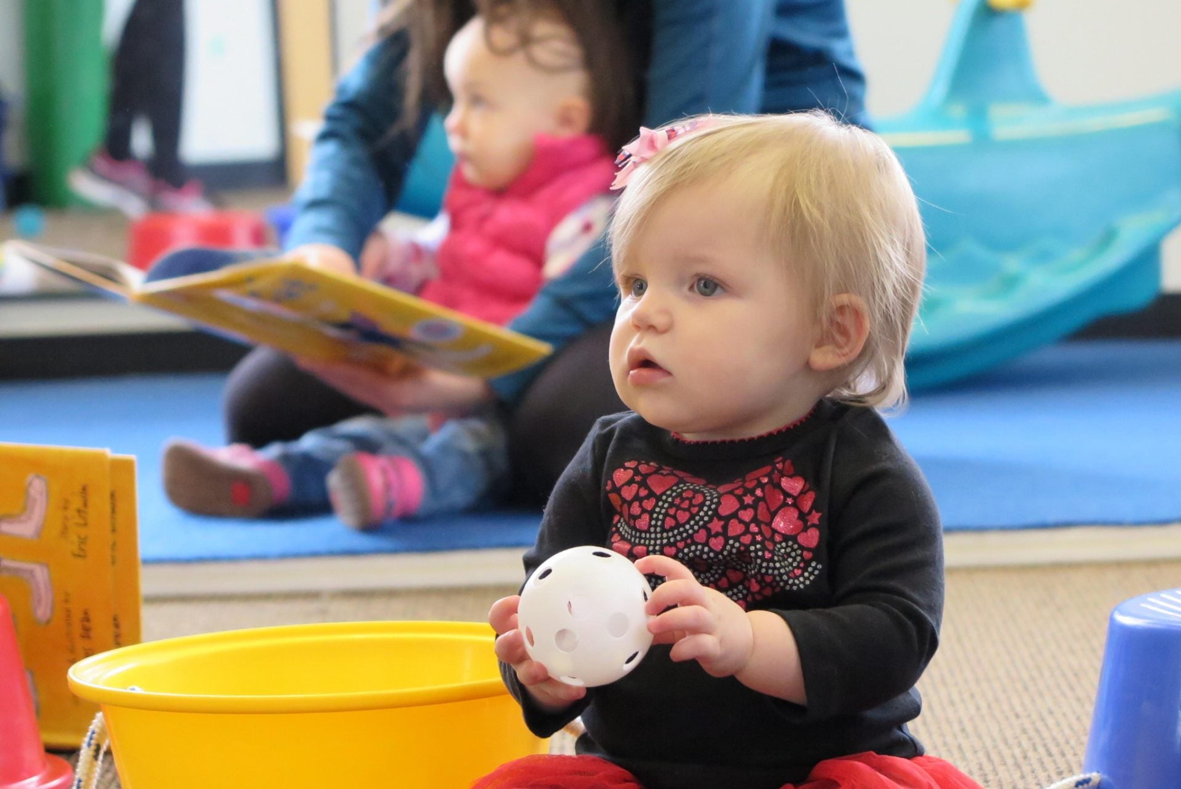 A toddler holds a white ball over a yellow bin while an adult reads a book to another child behind her