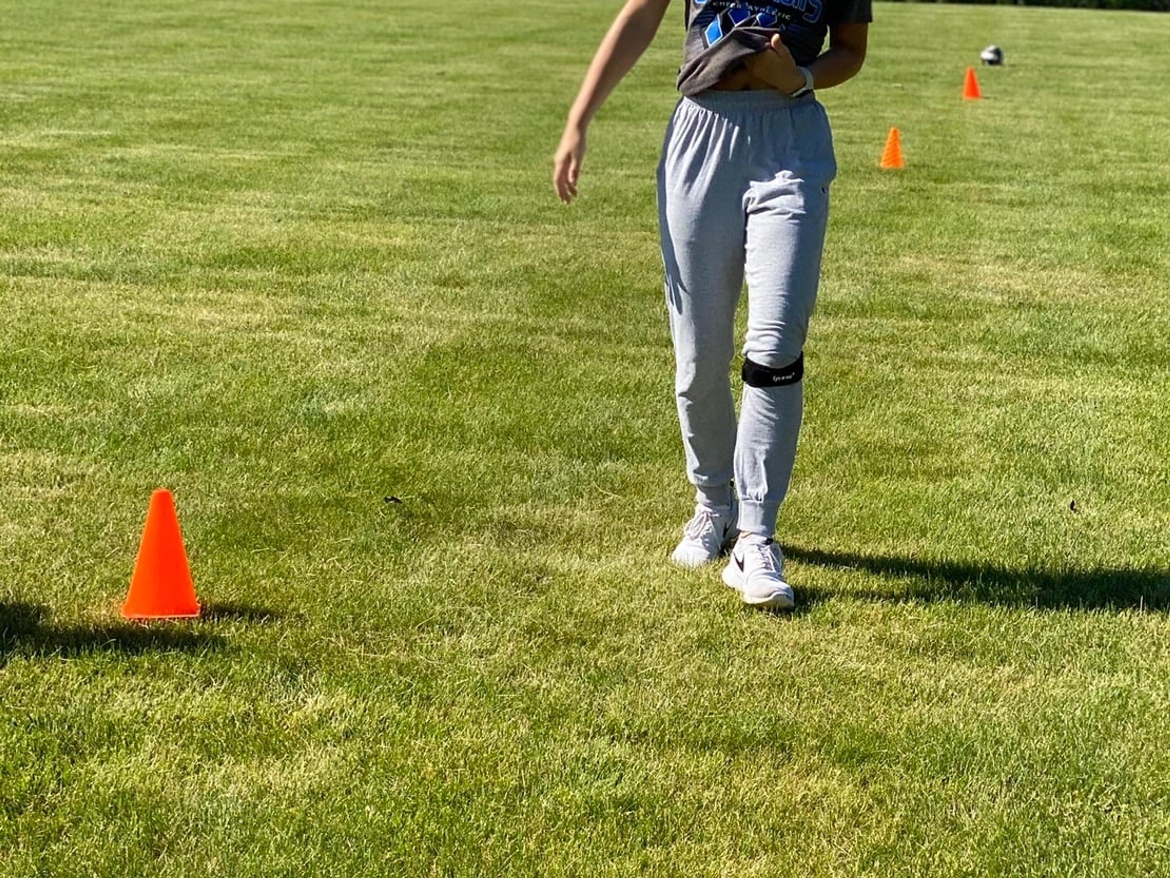 A smiling teen athlete wearing knee straps steps between orange cones on a grass field