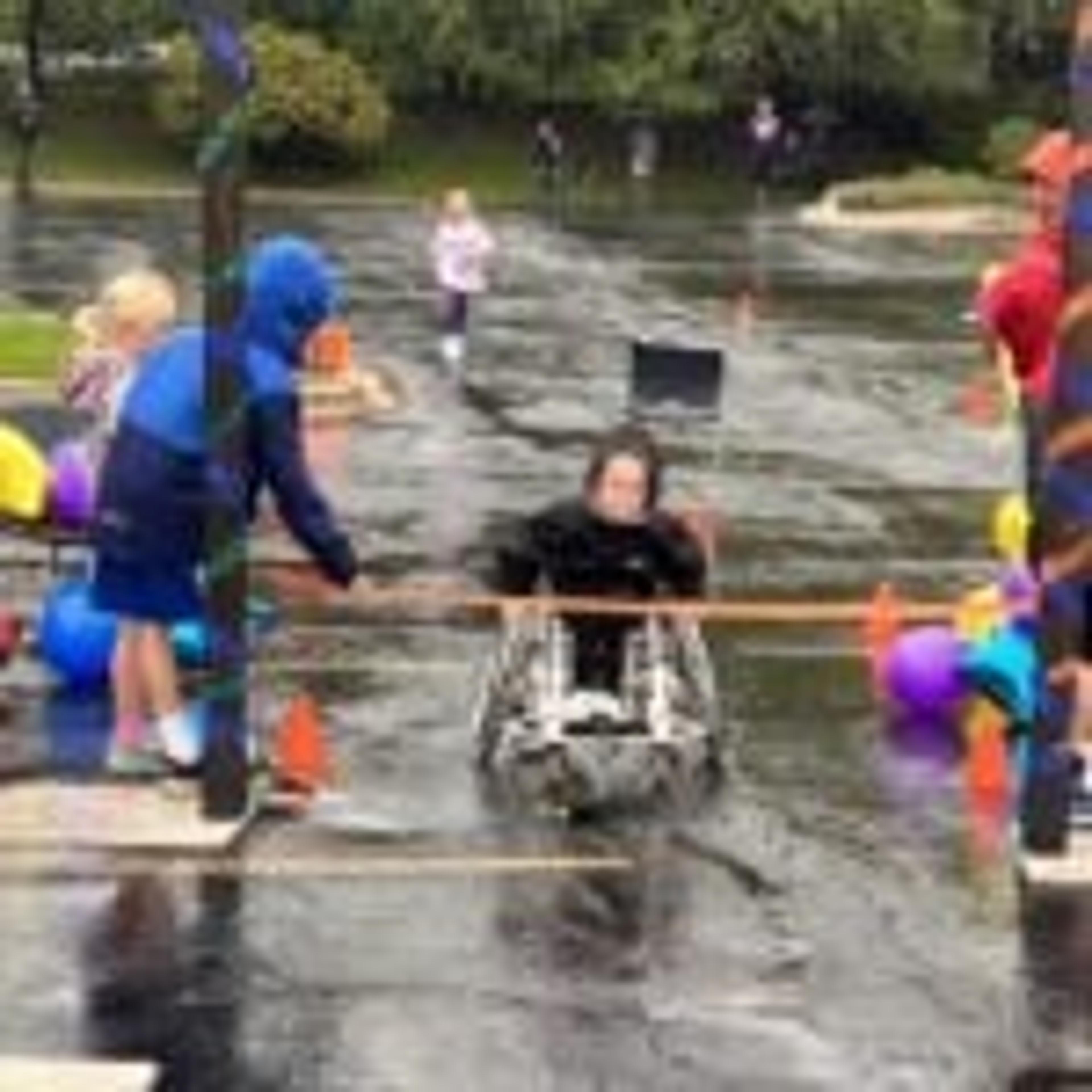 A participant in a wheeled mobility device heads toward a finish ribbon held by volunteers in rain jackets