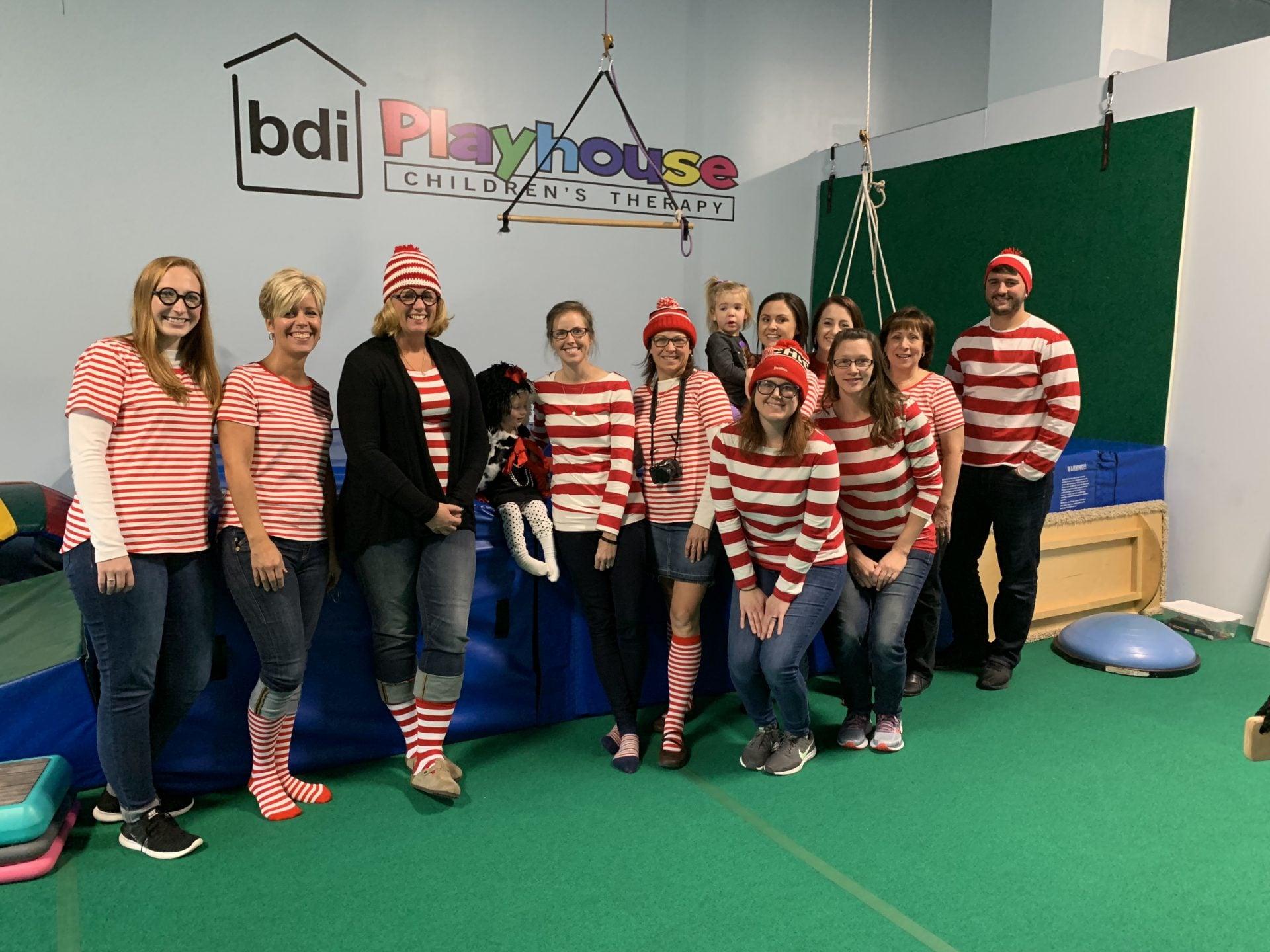Staff dressed in red-and-white striped Where's Waldo costumes pose in the gym beneath the BDI Playhouse sign