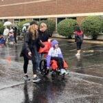 An adult pushes a smiling child in a wheelchair along the rain-soaked course as others walk behind