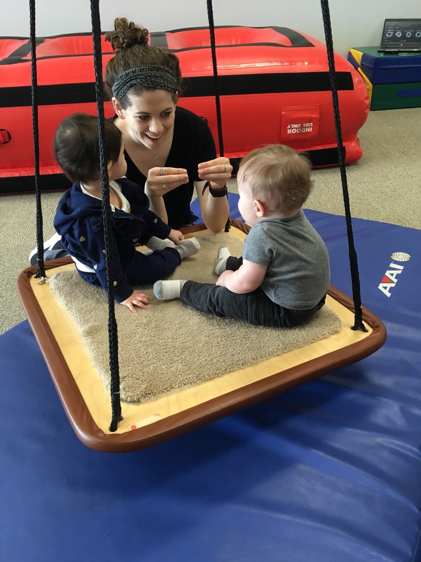 A therapist shows a hand sign to two babies sitting on a platform swing
