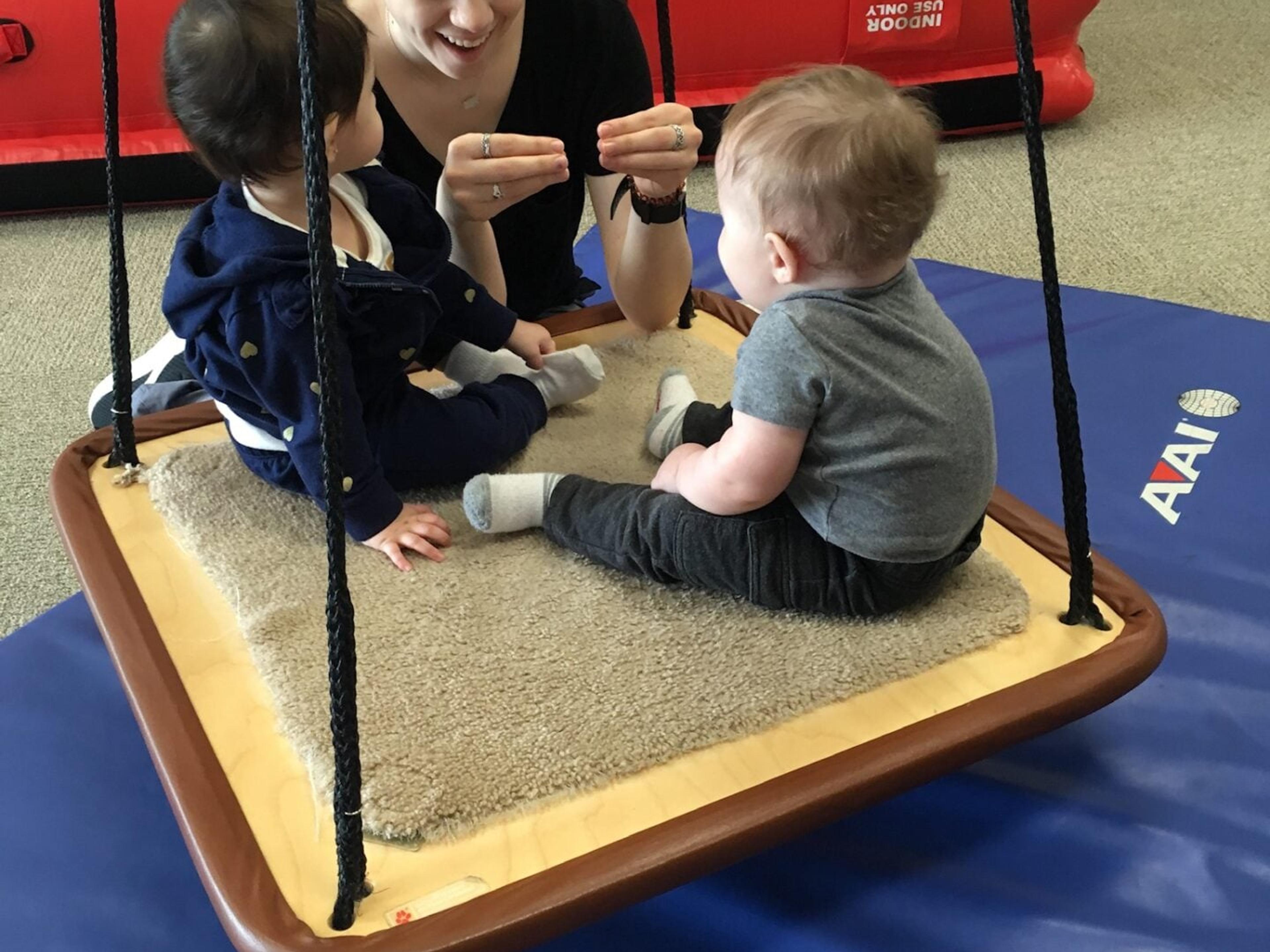 A therapist shows a hand sign to two babies sitting on a platform swing