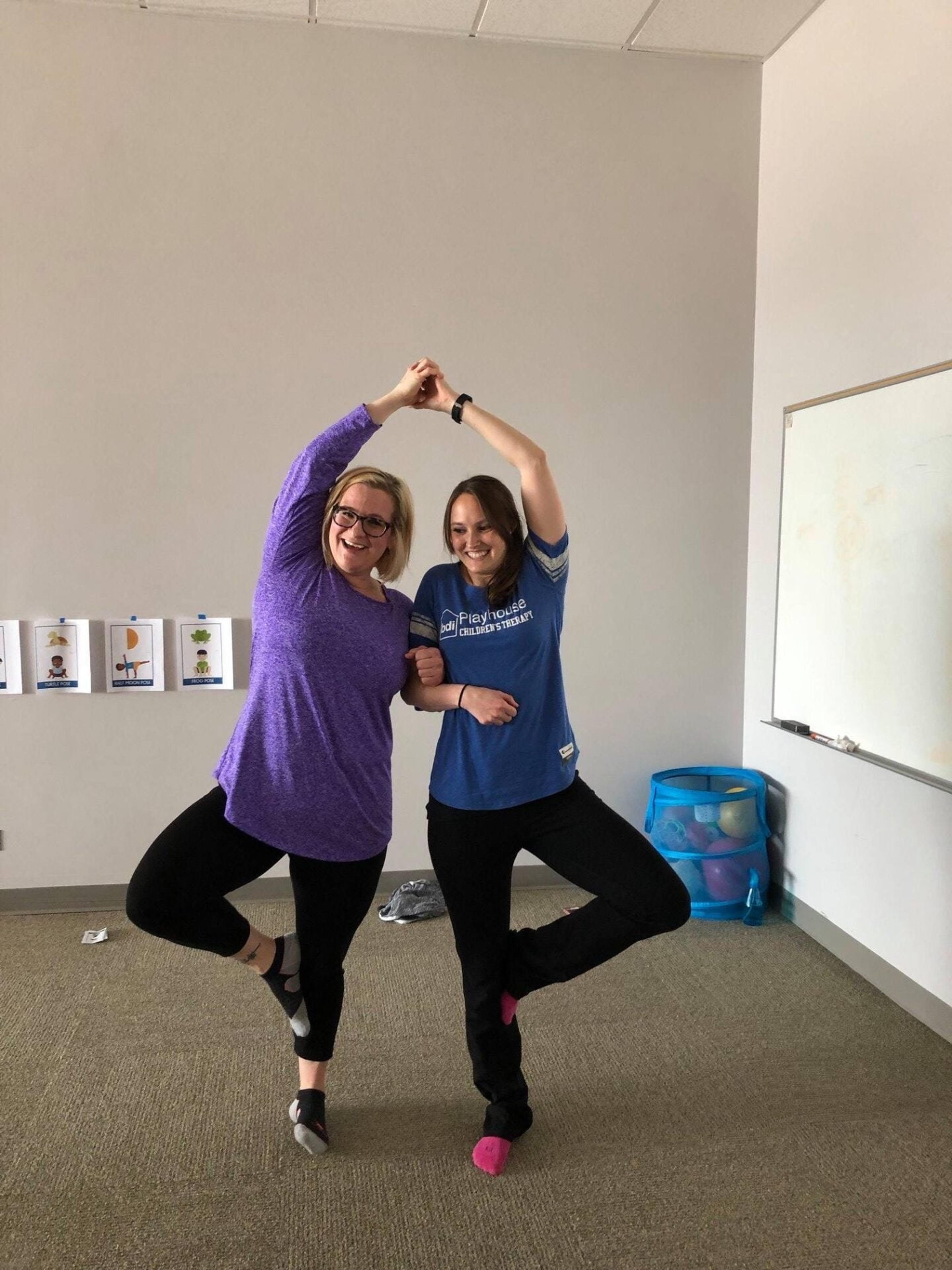 Two staff members balance on one leg with hands joined overhead in a playful pose in a meeting room