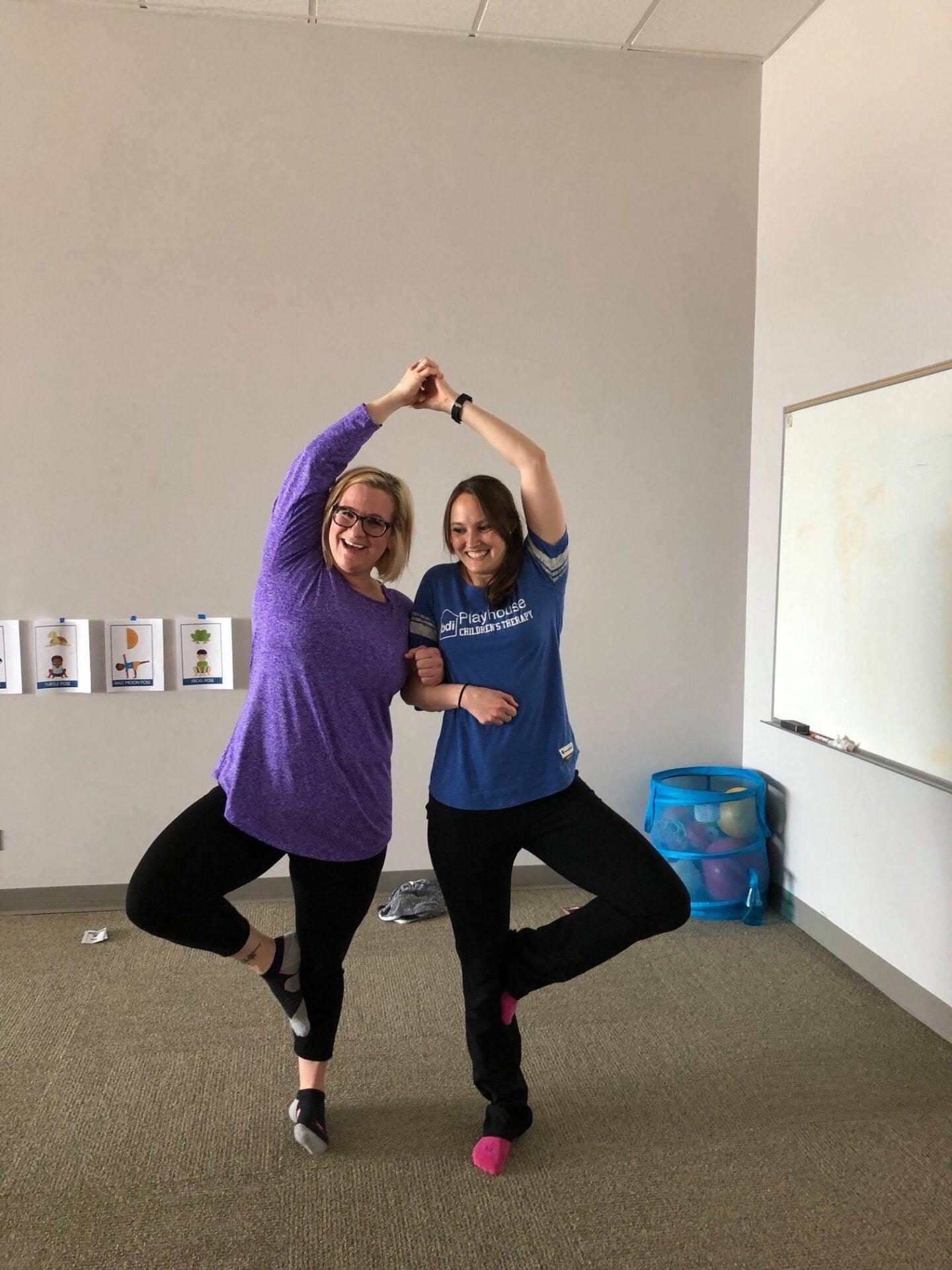 Two staff members balance on one leg with hands joined overhead in a playful pose in a meeting room