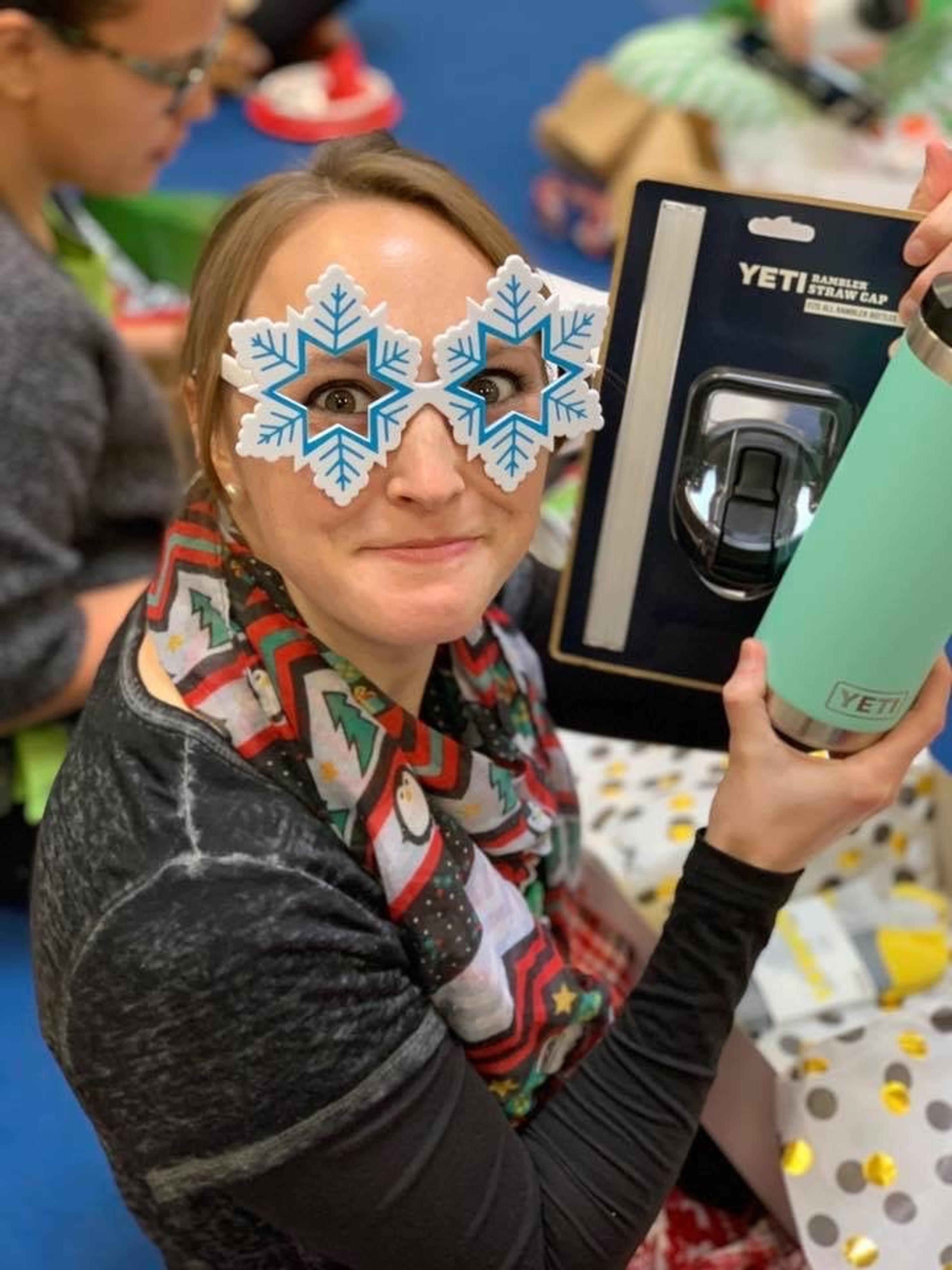 A staff member wearing snowflake-shaped glasses holds up a Yeti tumbler gift at a holiday party