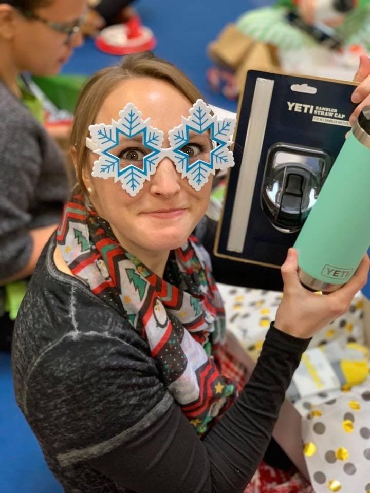 A staff member wearing snowflake-shaped glasses holds up a Yeti tumbler gift at a holiday party