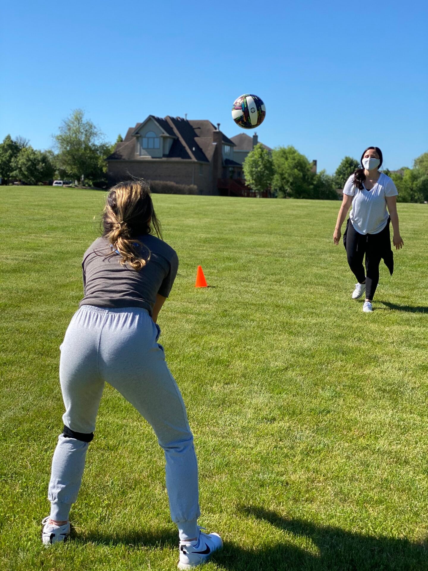 A teen athlete wearing knee straps gets set to play a ball tossed by a therapist in a face mask on a grass field