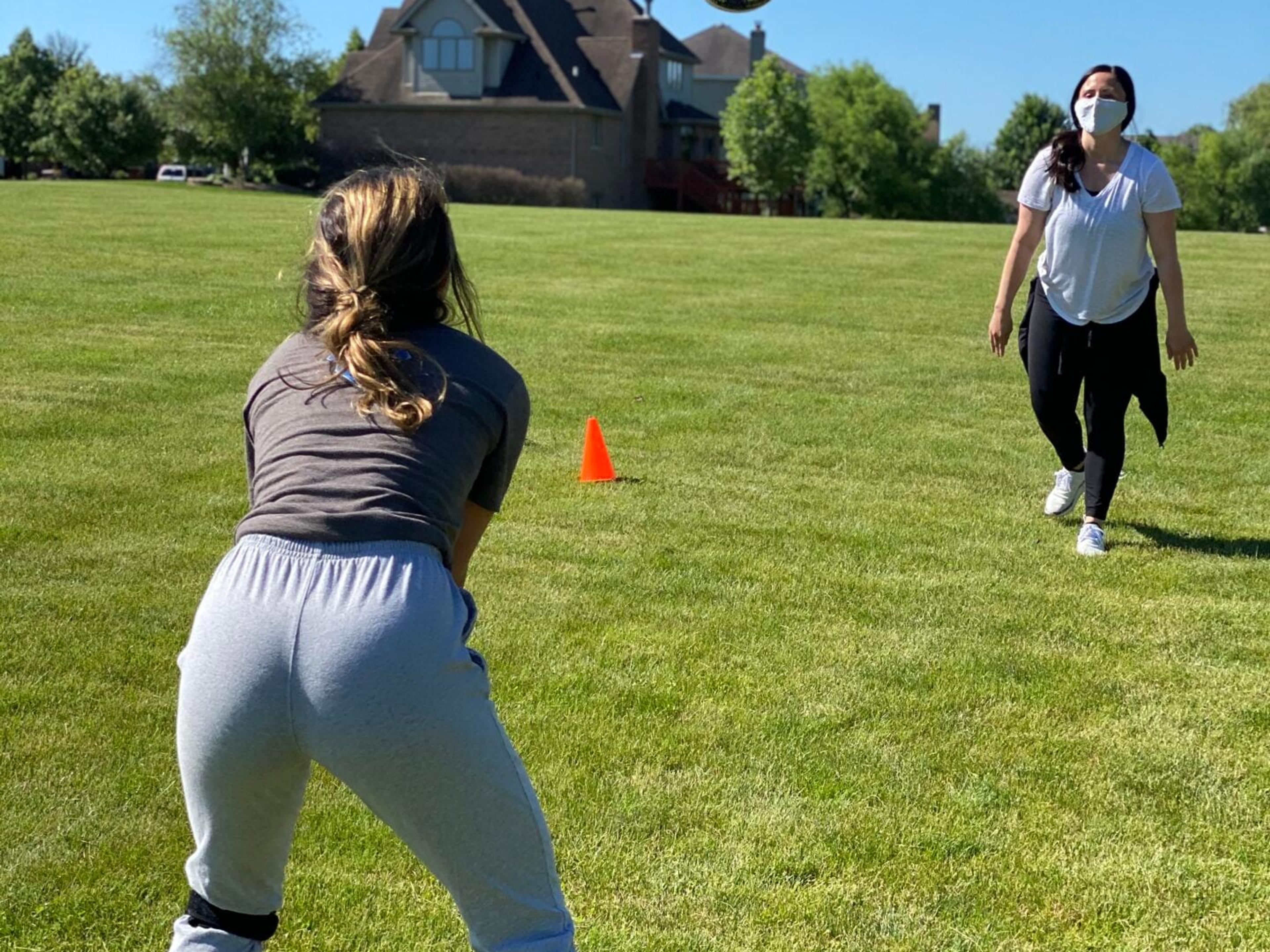A teen athlete wearing knee straps gets set to play a ball tossed by a therapist in a face mask on a grass field