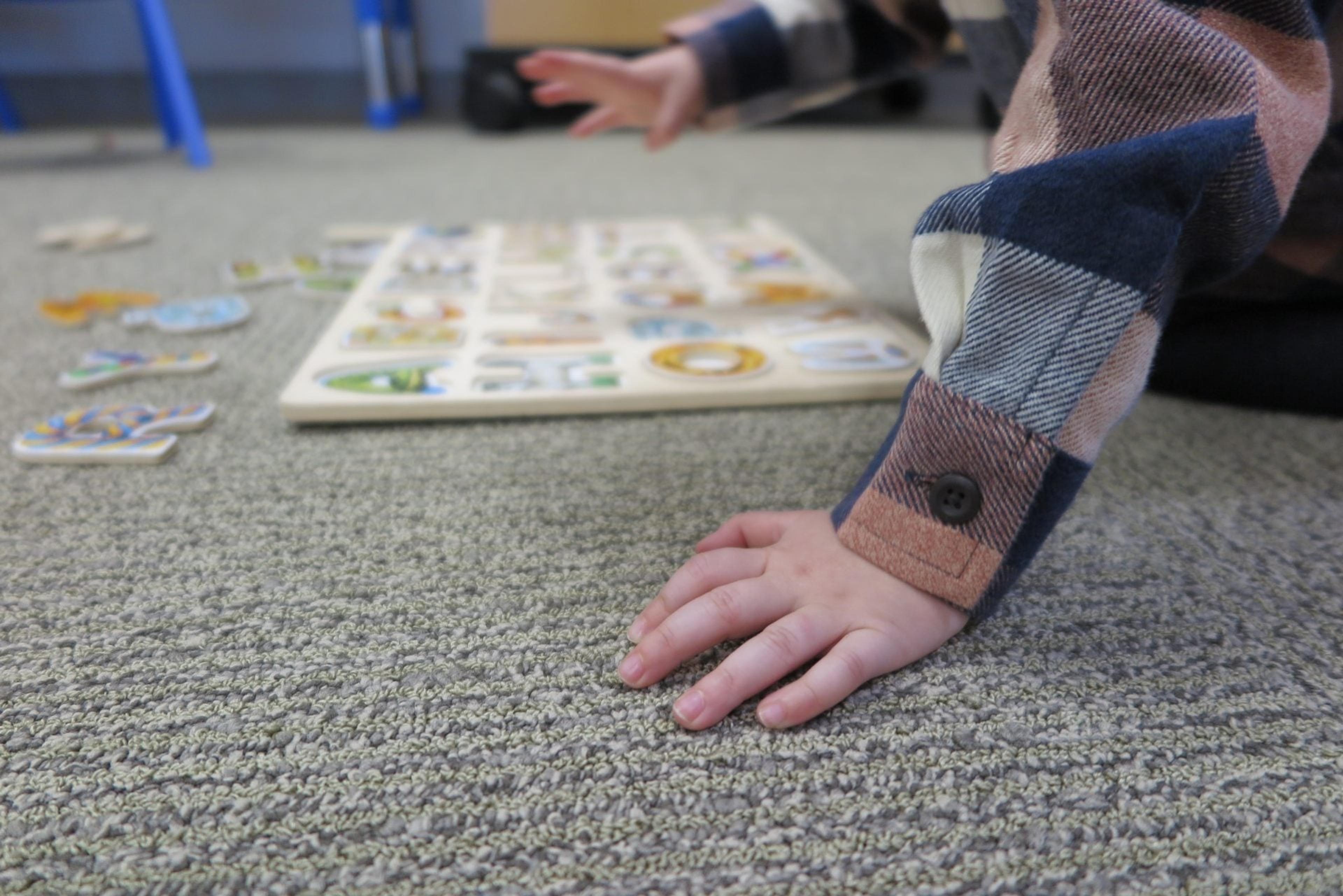 Close-up of a child's hand planted on the carpet while reaching toward a wooden alphabet puzzle