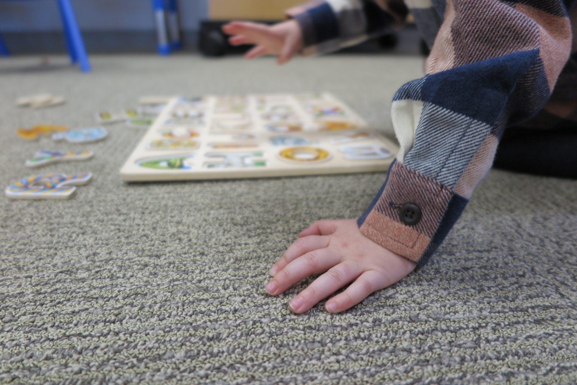 Close-up of a child's hand planted on the carpet while reaching toward a wooden alphabet puzzle