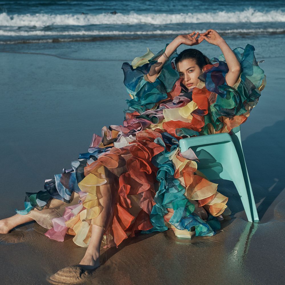 Model sitting in chair on beach in ruffled gown. 