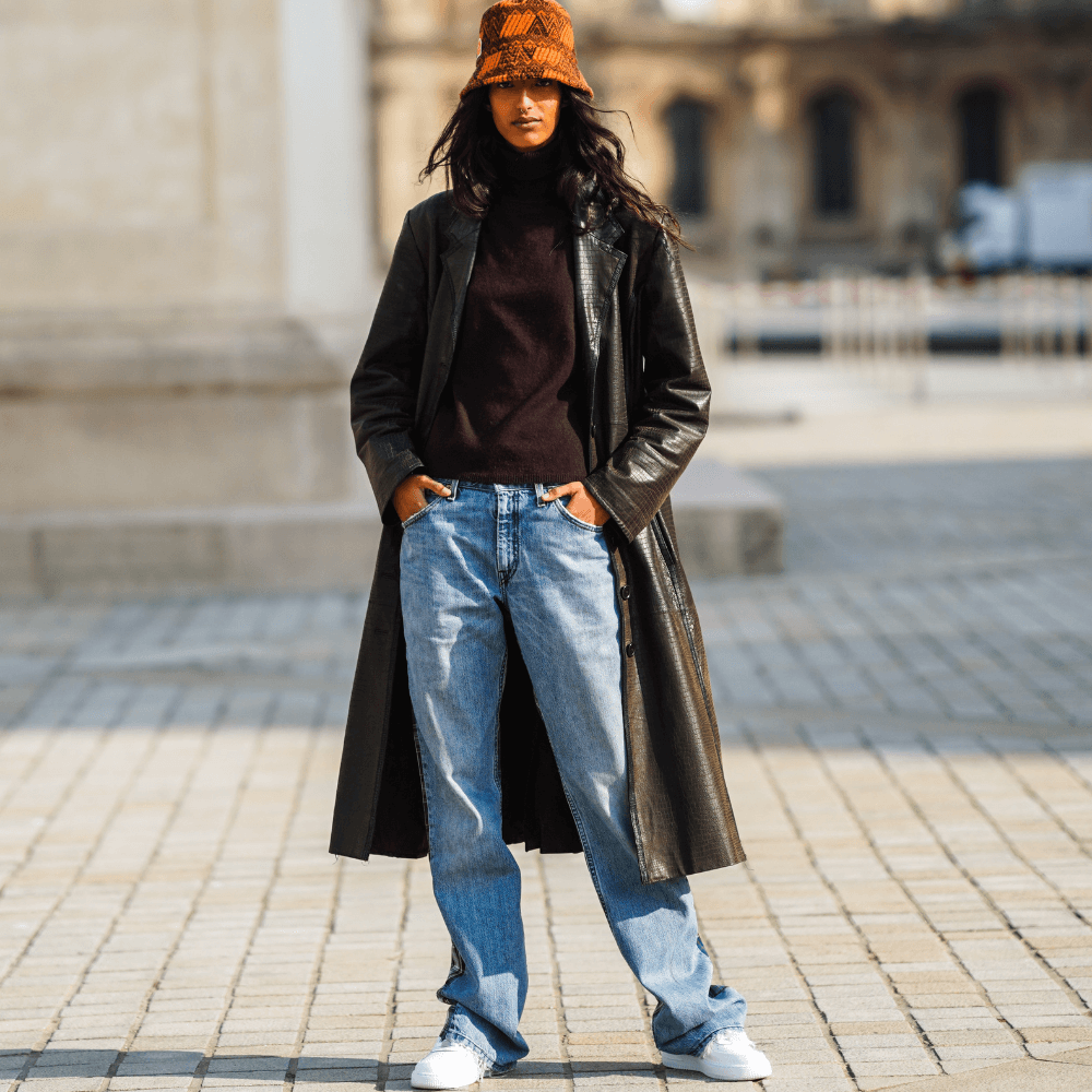 A model wears a brown and orange bob hat from Prada, a brown leather long trench coat with crocodile pattern, a burgundy wool pullover, blue denim jeans pants with checked printed parts, white sneakers