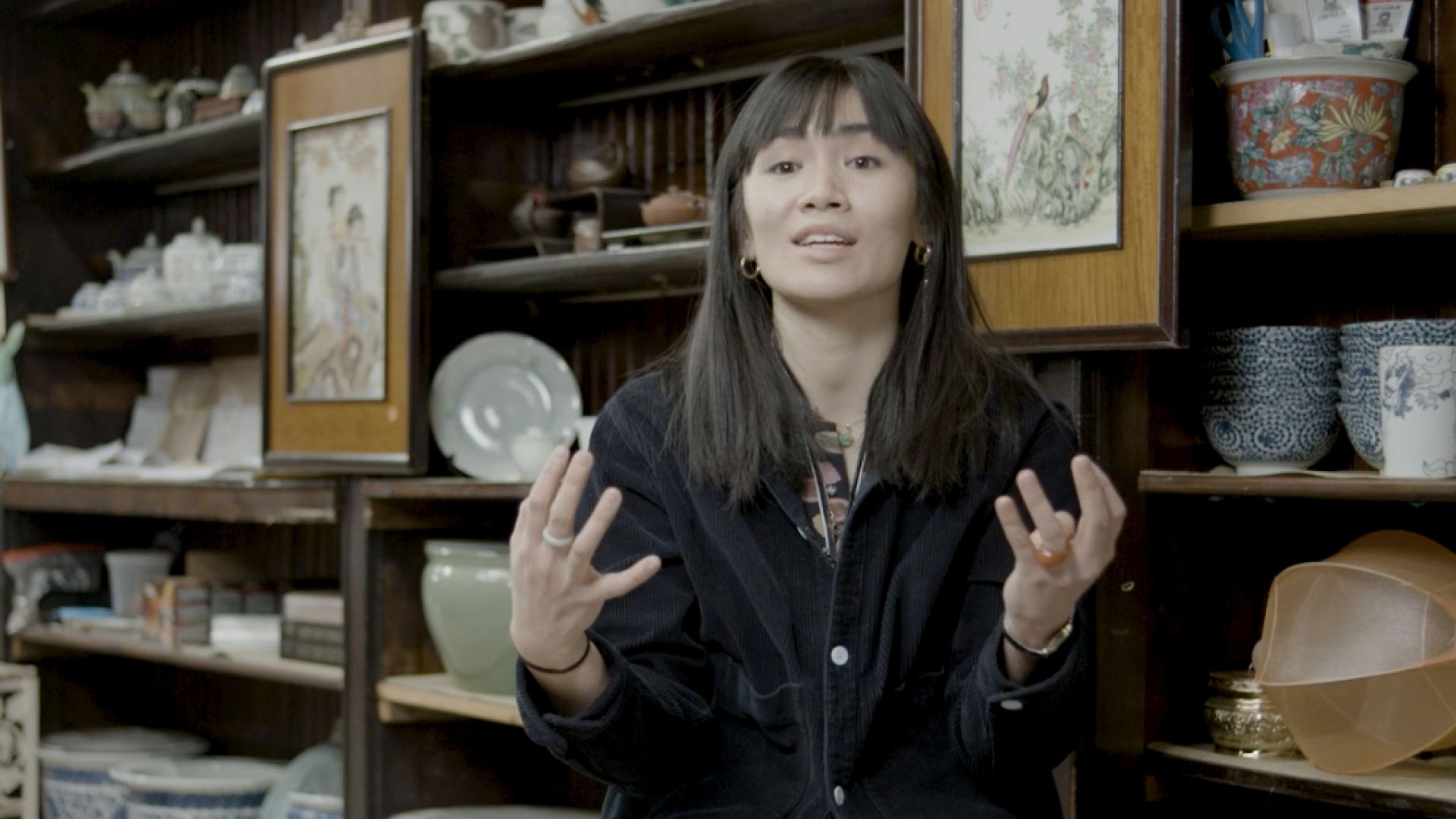Artist Mei Lum gestures with her hands as she speaks at Wing on Wo store in front of porcelain ware home items.
