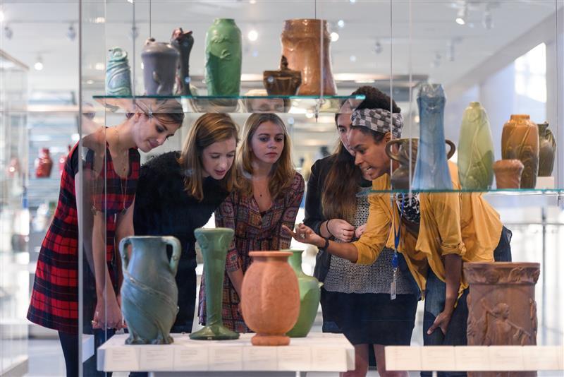 A small group of young people talking in front of a multi-tier glass case full of blue, green, and tan vases and pots.