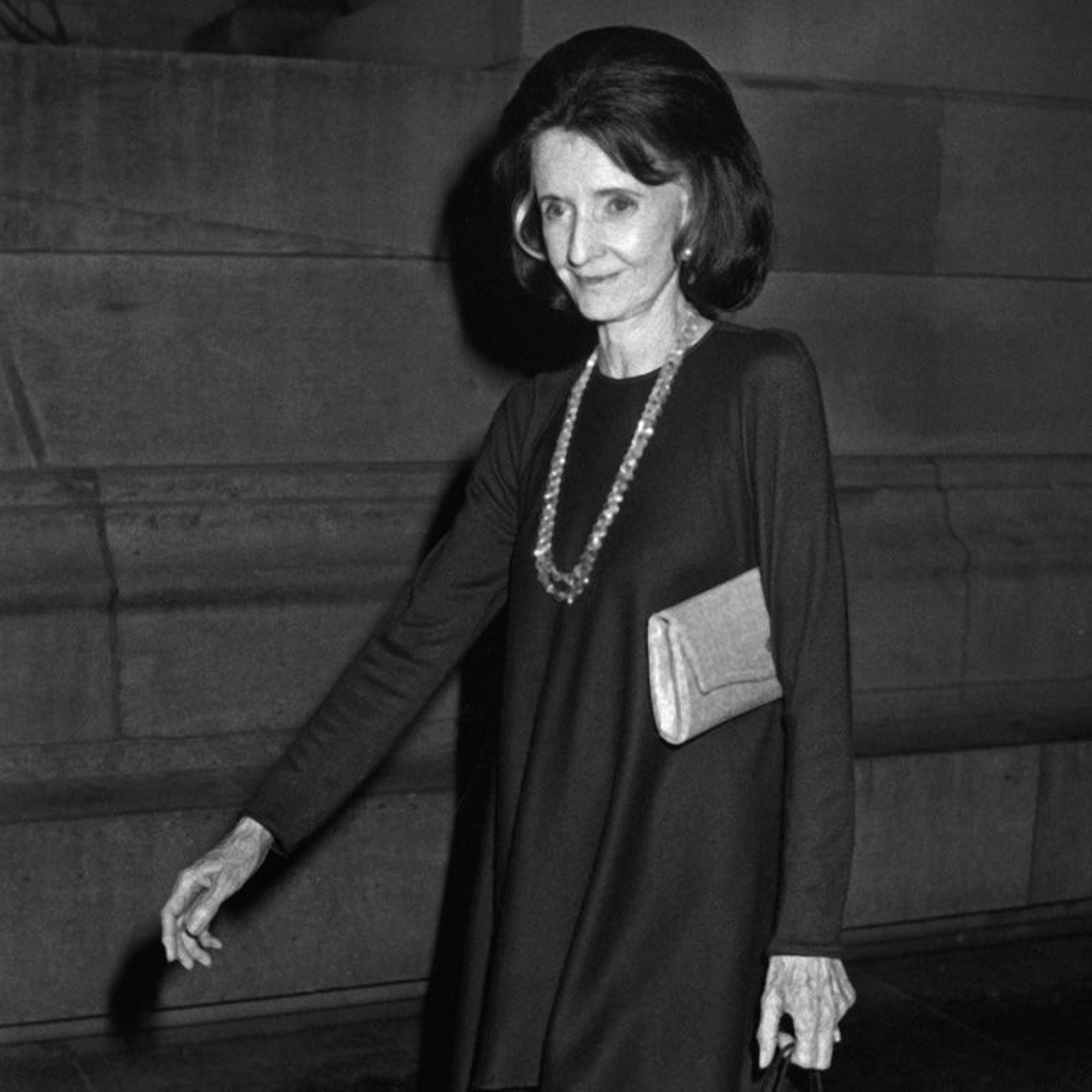 A stylish woman in a long black dress and a statement necklace, holding a small clutch, walking against a textured background.
